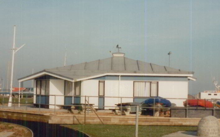 A small, white, octagonal building with a metal roof, surrounded by a wooden deck with picnic tables and a few parked cars nearby.