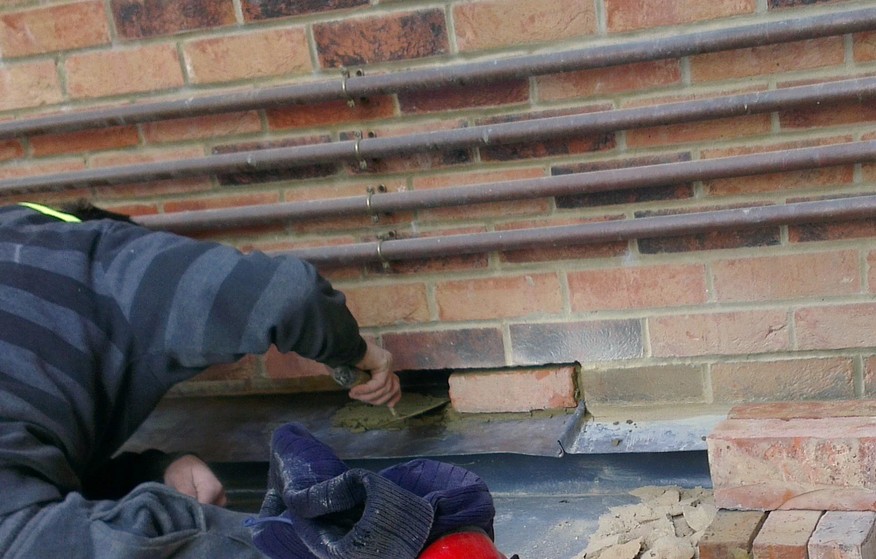 A worker laying bricks and applying mortar to a brick wall with metal pipes mounted horizontally.