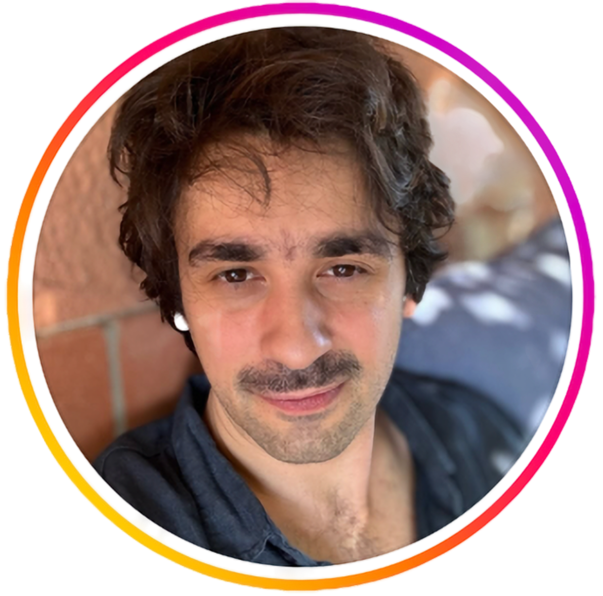Close-up of a young man with wavy dark hair, brown eyes, light facial hair, smiling slightly, wearing a dark shirt.