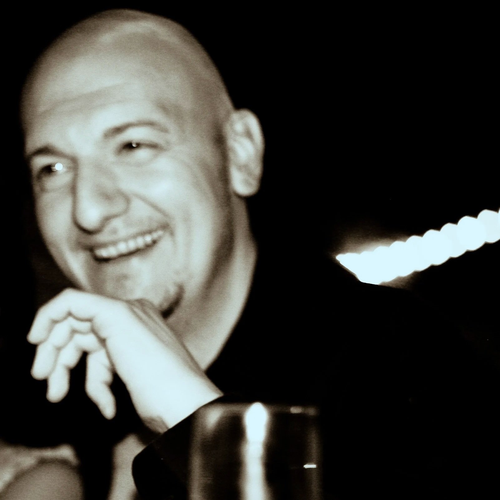 Black and white photo of a smiling man with short hair, resting his chin on his hand, with a blurred background.