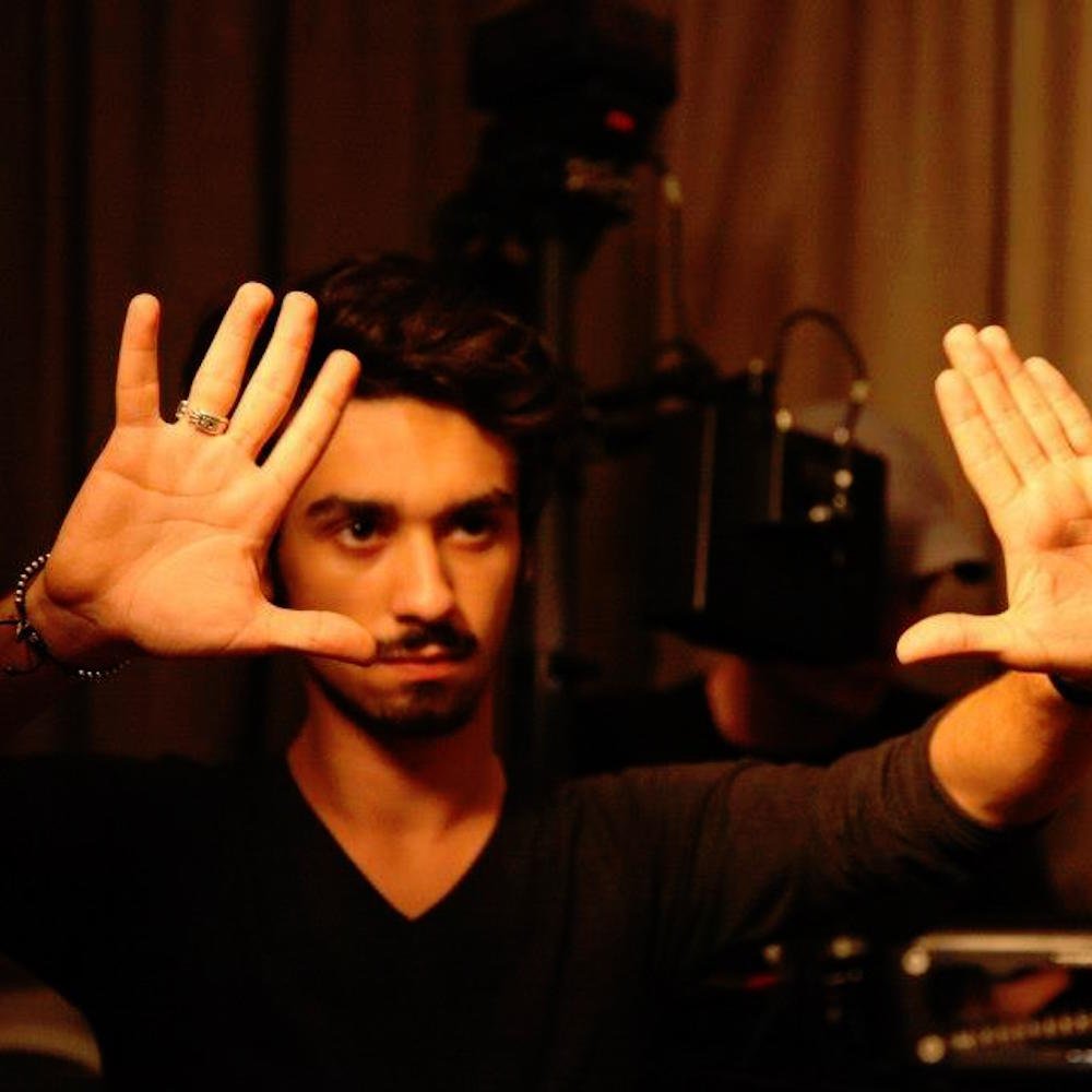 A filmmaker with dark hair and a beard holds both of his hands up, palms facing outward, in front of a camera. He is wearing a black shirt and some jewelry on his fingers and wrist.