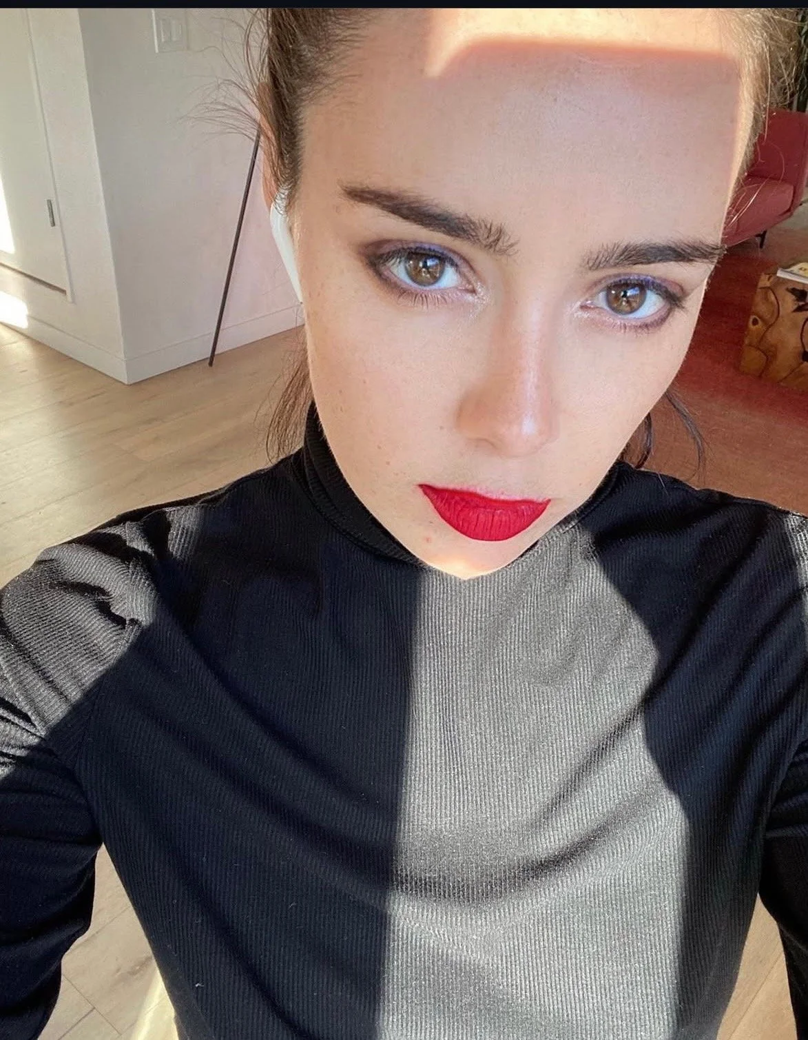 A woman with blue eyes and red lipstick taking a selfie indoors in natural light, wearing a black top with textured fabric.