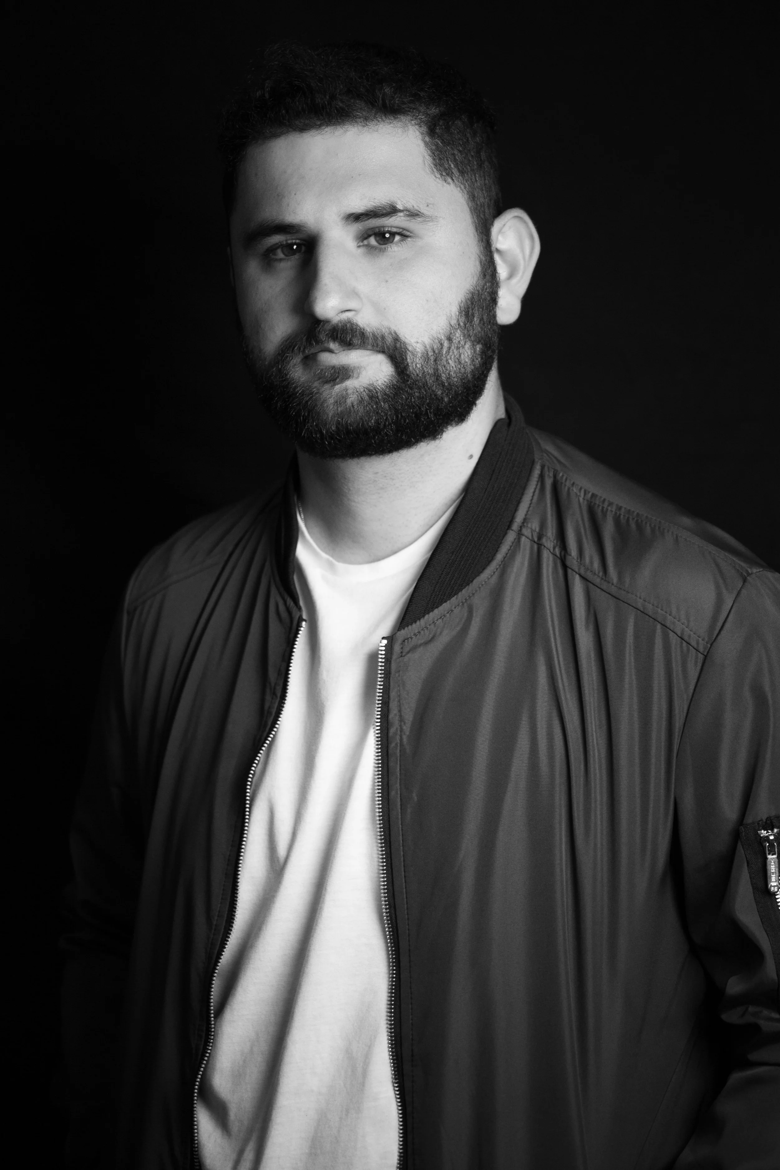 Black and white portrait of a man with a beard and short hair, wearing a zip-up jacket over a white t-shirt, against a dark background.