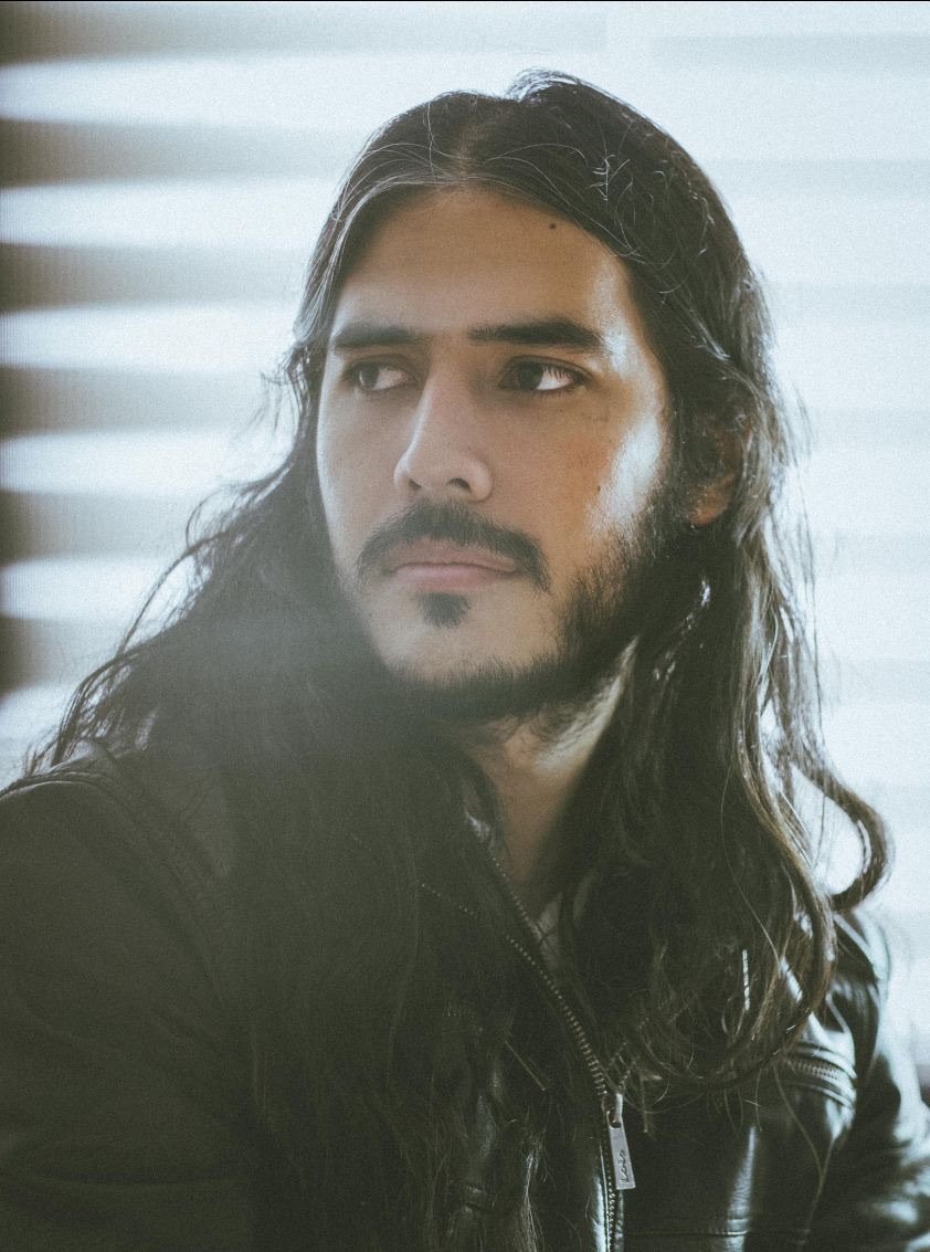 A man with long, dark hair and a beard, wearing a black leather jacket, looking to his left indoors.