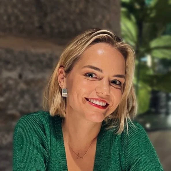 A woman with blonde hair smiling, wearing a green sweater and square earrings, in an outdoor setting with a stone and green foliage background.