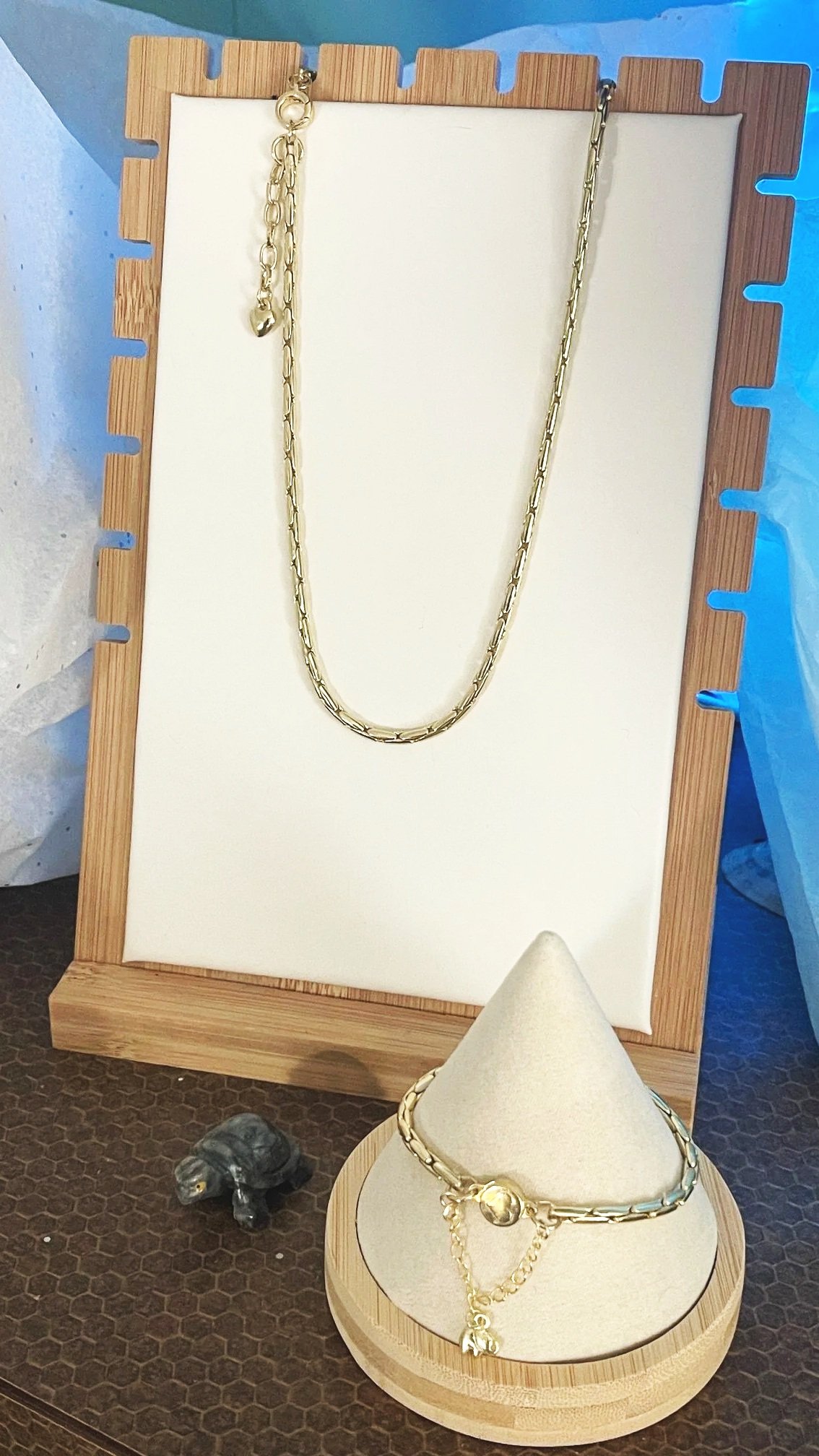 Gold jewelry displayed on a wooden and white jewelry stand, including a chain necklace and matching bracelet with a charm. There is also a small black decorative elephant figurine nearby.