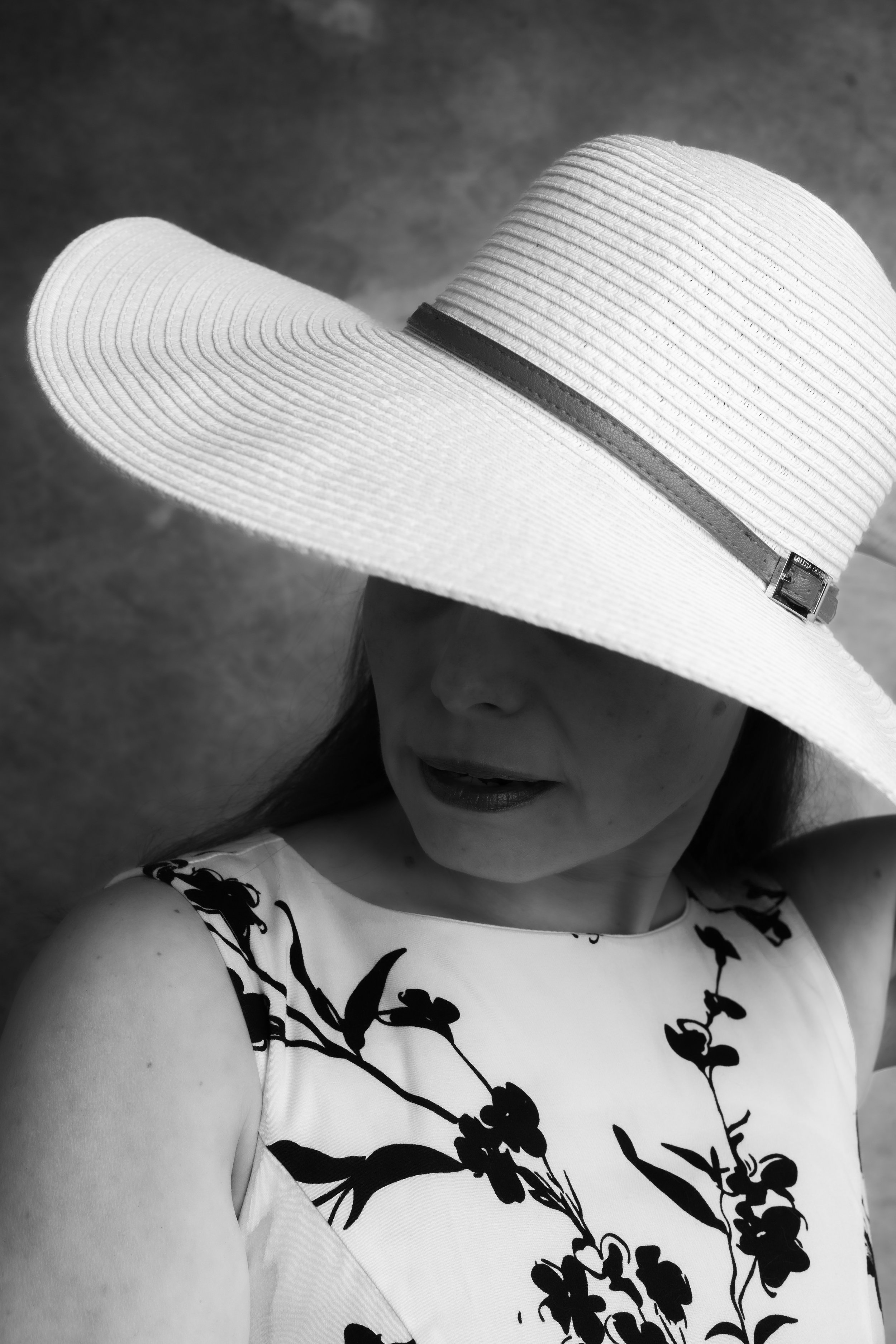 A woman wearing a wide-brimmed straw hat with a dark band and a sleeveless dress with a black floral pattern, partially visible in a black-and-white photograph.