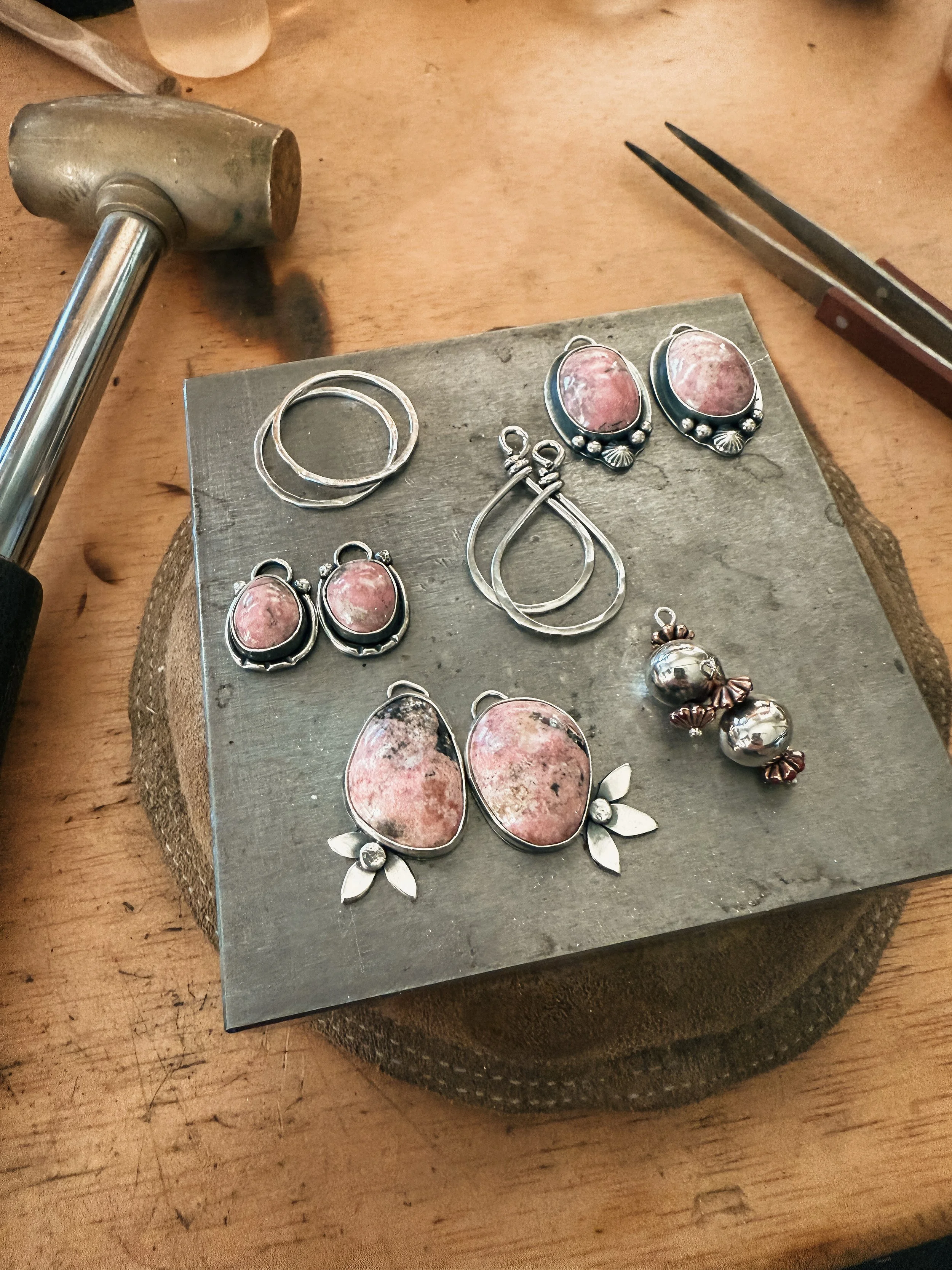 Silver jewelry with pink stones, including earrings and pendants, arranged on a metal board, surrounded by jewelry-making tools on a wooden workbench.