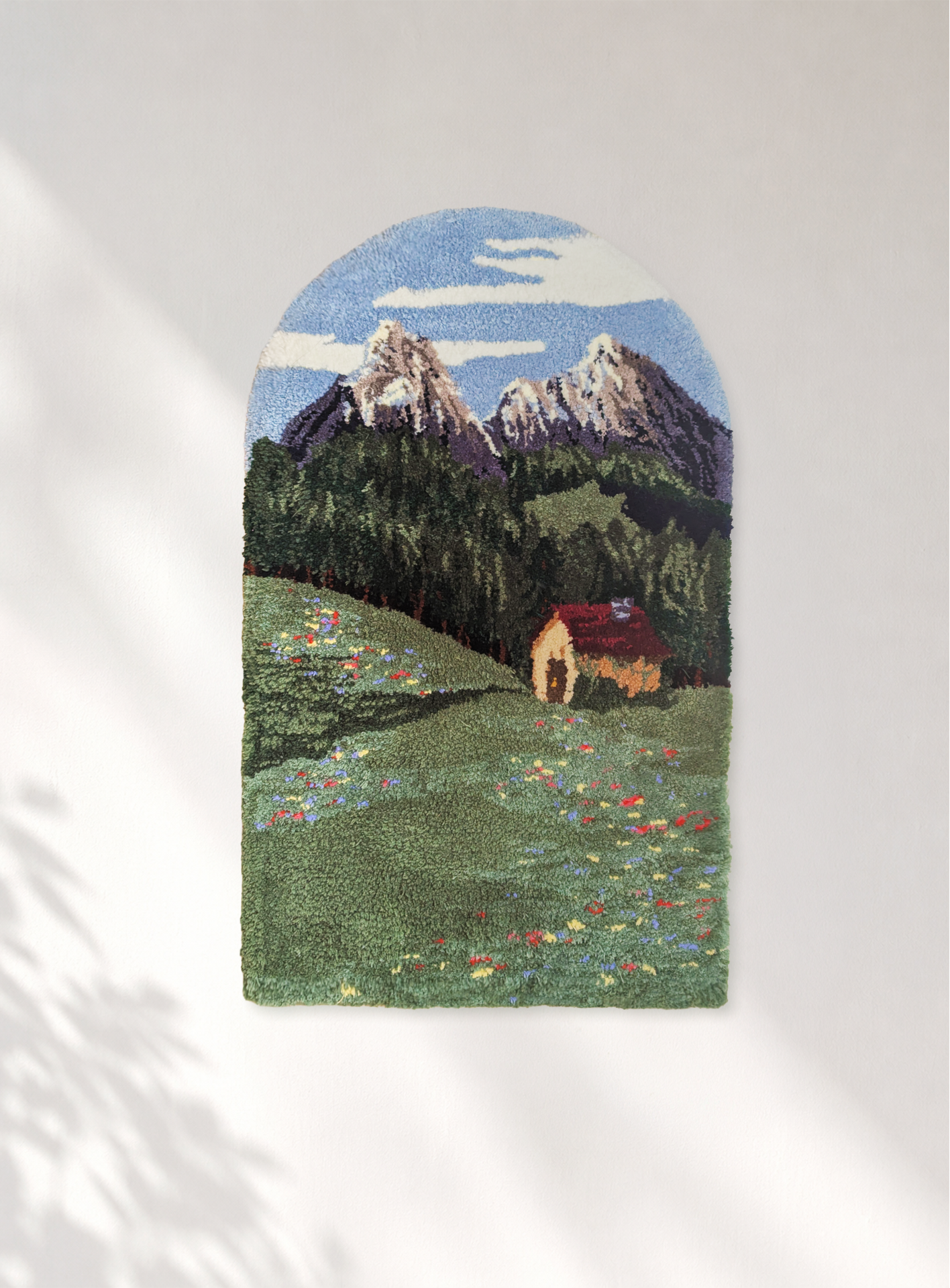 Handmade tufted landscape featuring snow-capped mountains, pine trees, a grassy meadow with colorful wildflowers, and a small cottage with a red roof, under a blue sky with clouds.