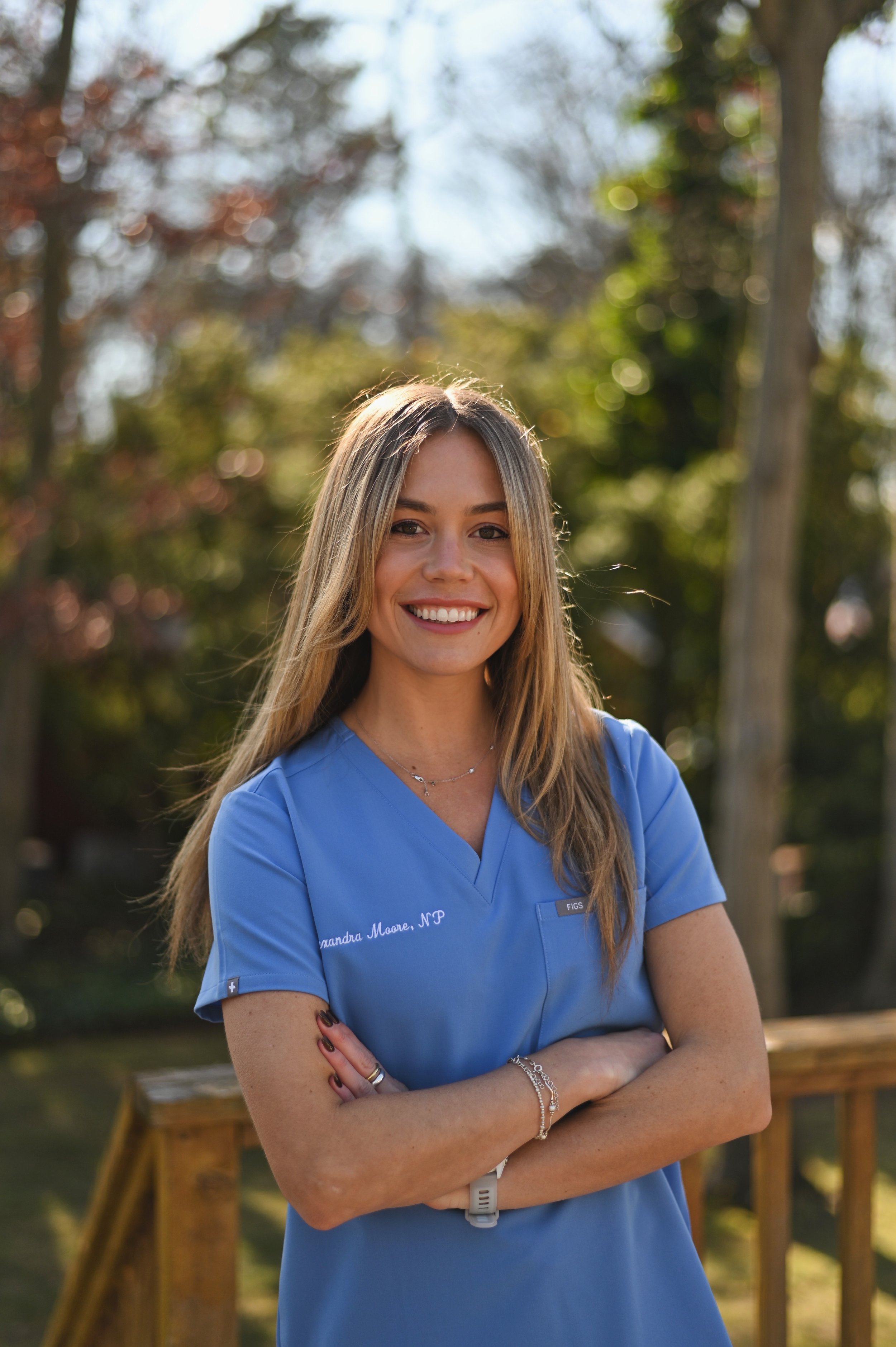 A young female nurse practitioner with long blonde hair smiling outdoors, wearing blue medical scrubs with her arms crossed.