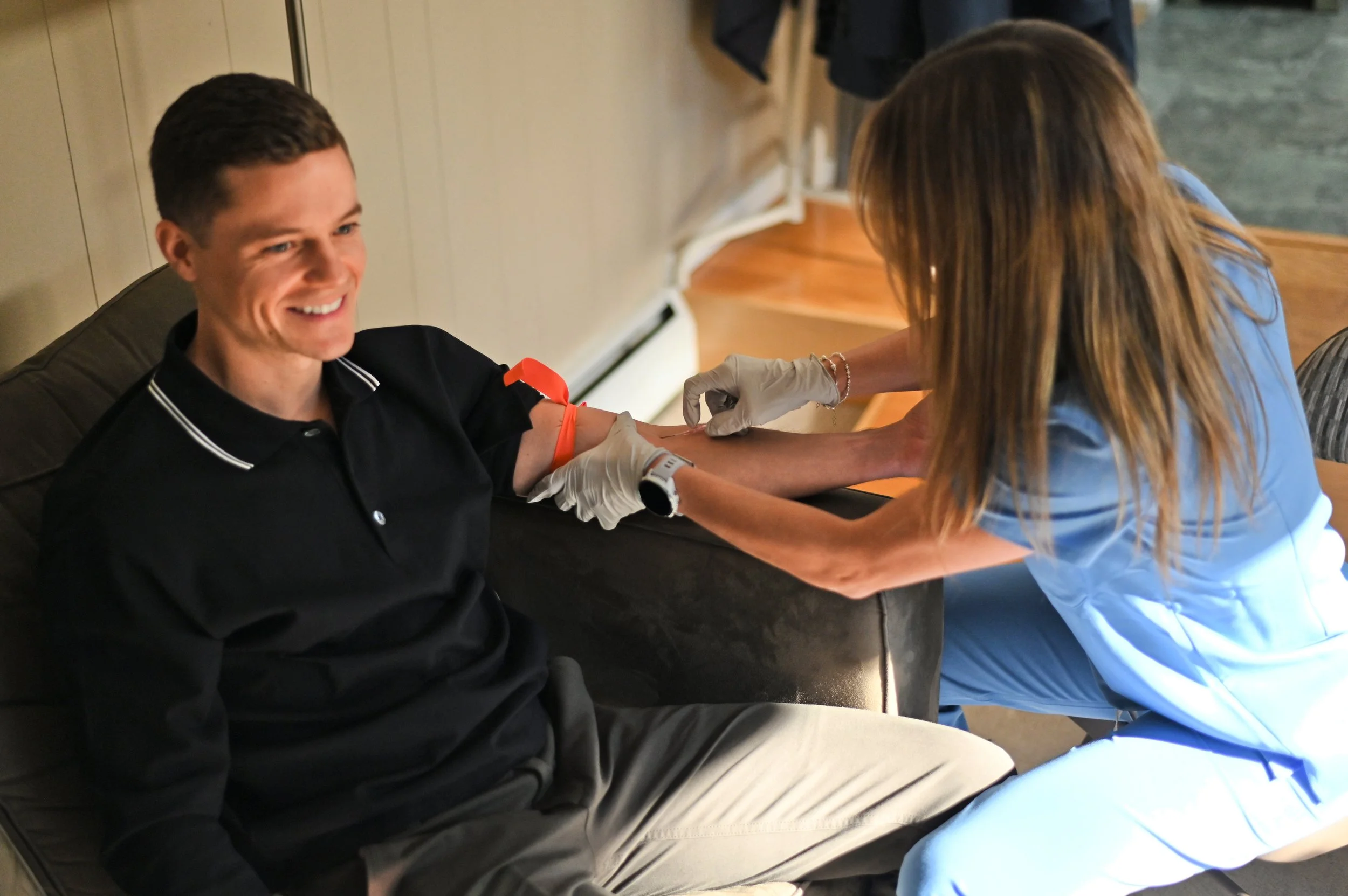A man sitting on a chair smiling while a nurse practitioner gives him an IV in his left arm.