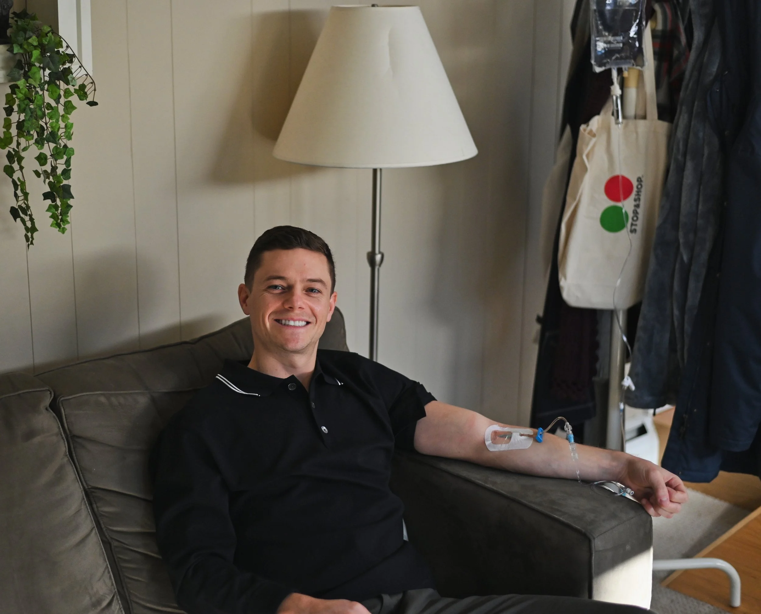 A smiling young man sitting on a beige sofa, getting an IV infusion with vitamins, in a room with a floor lamp, hanging plants, and a coat rack with bags and jackets.