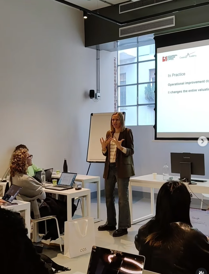 Une femme présente en face d'un groupe lors d'une séance de formation ou de conférence, avec un écran et un tableau blanc dans une salle de réunion.