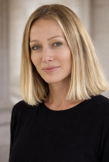 Portrait d'une femme blonde avec une expression neutre, portant un pull noir, à l'intérieur.