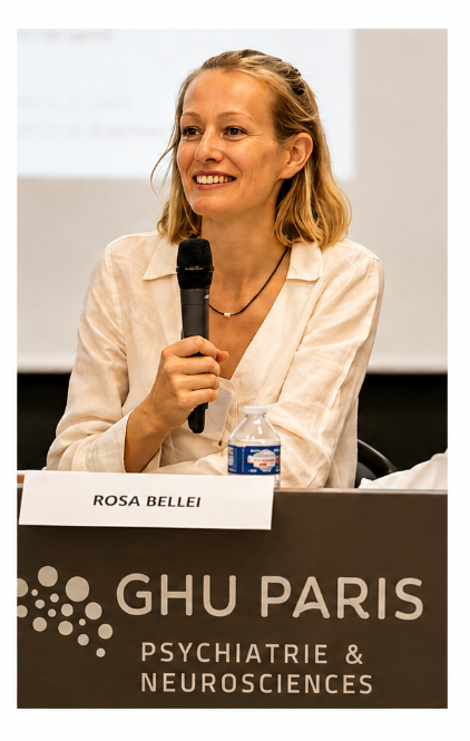 Une femme souriante avec des cheveux blonds courts, portant une chemise beige clair, tient un micro lors d'une conférence. Sur la table, il y a un nom porté 'ROSA BELLEI', une bouteille d'eau en plastique. Le panneau indique 'GHU PARIS PSYCHIATRIE & NEUROSCIENCES'.