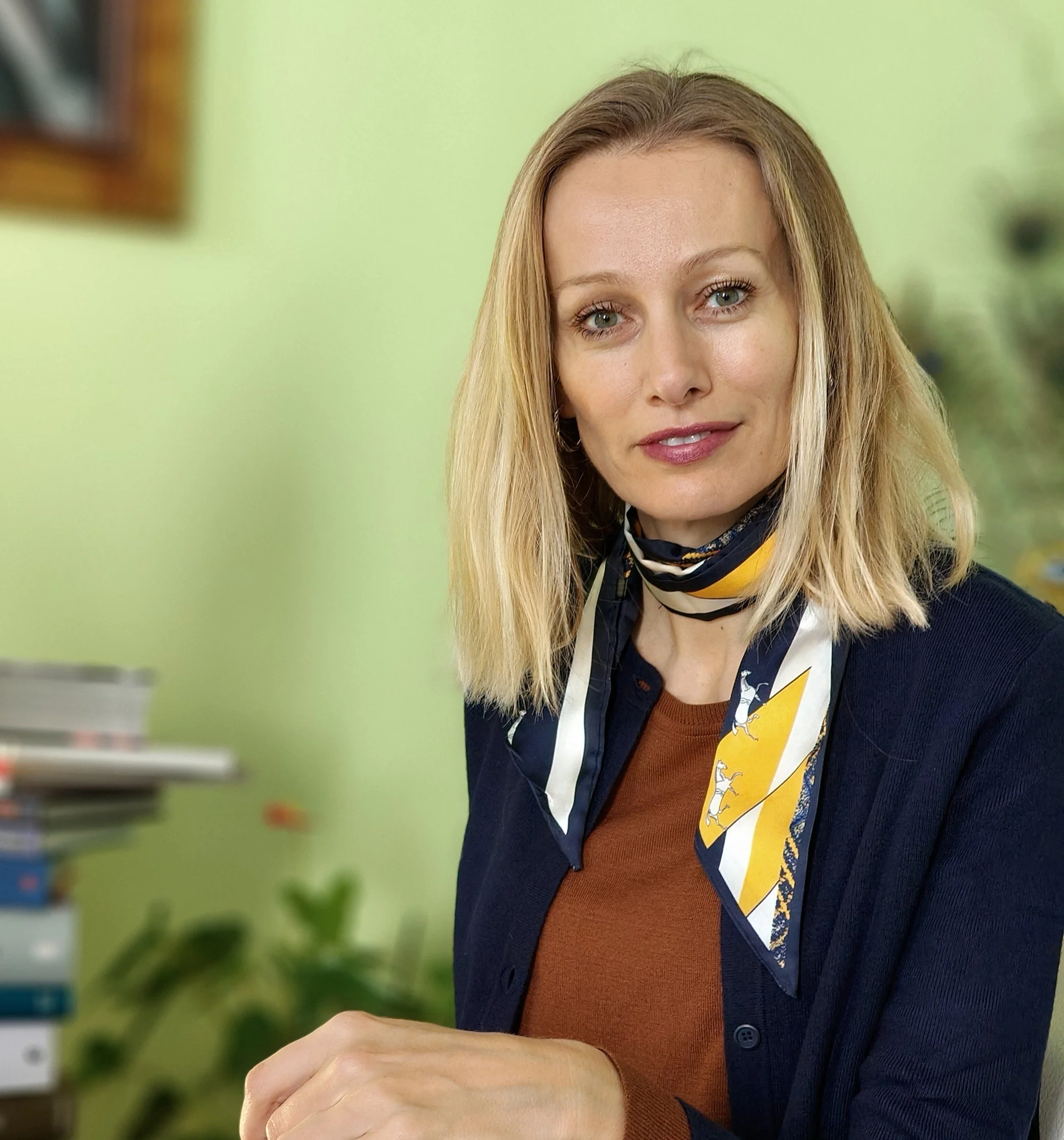 Une femme blonde portant une veste noire et un foulard avec une cravate jaune, assise à une table dans un environnement intérieur avec un mur vert en arrière-plan.