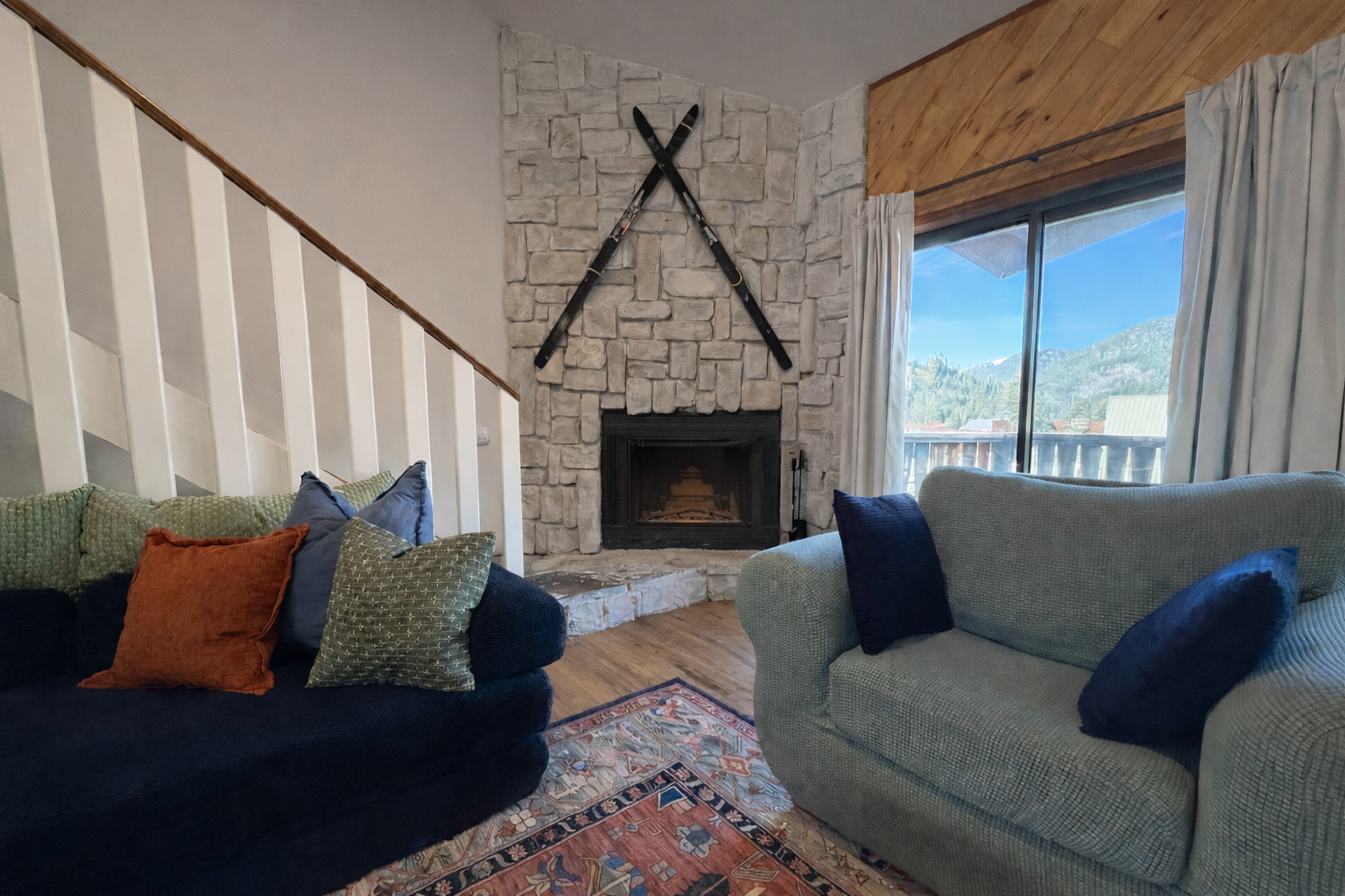 Living room with a stone fireplace, two crossed skis above it, large window with mountains view, two sofas with cushions, and a colorful rug.