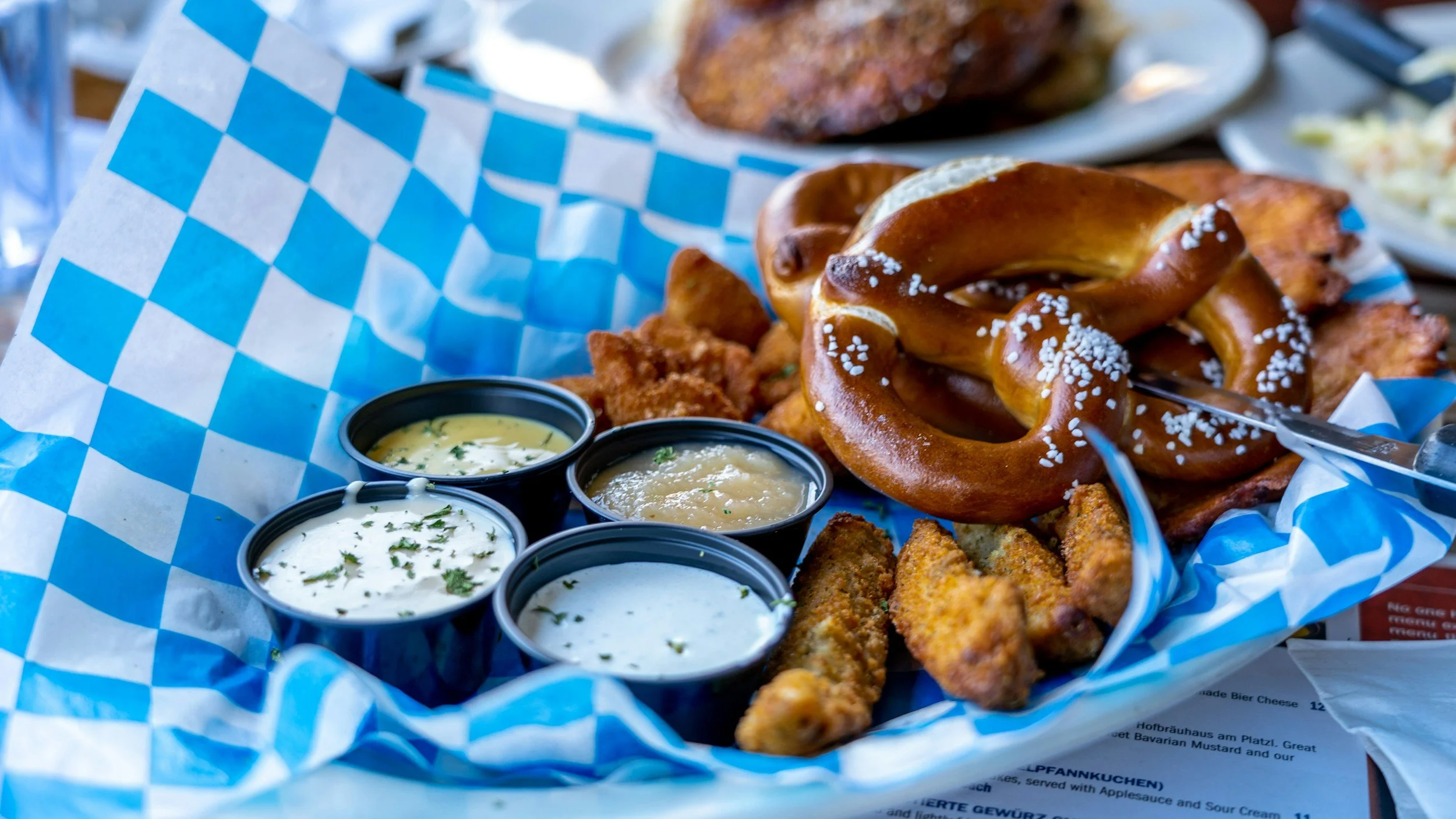 A basket of German-style pretzels with coarse salt, accompanied by small cups of dipping sauces, on a blue and white checkered paper liner.