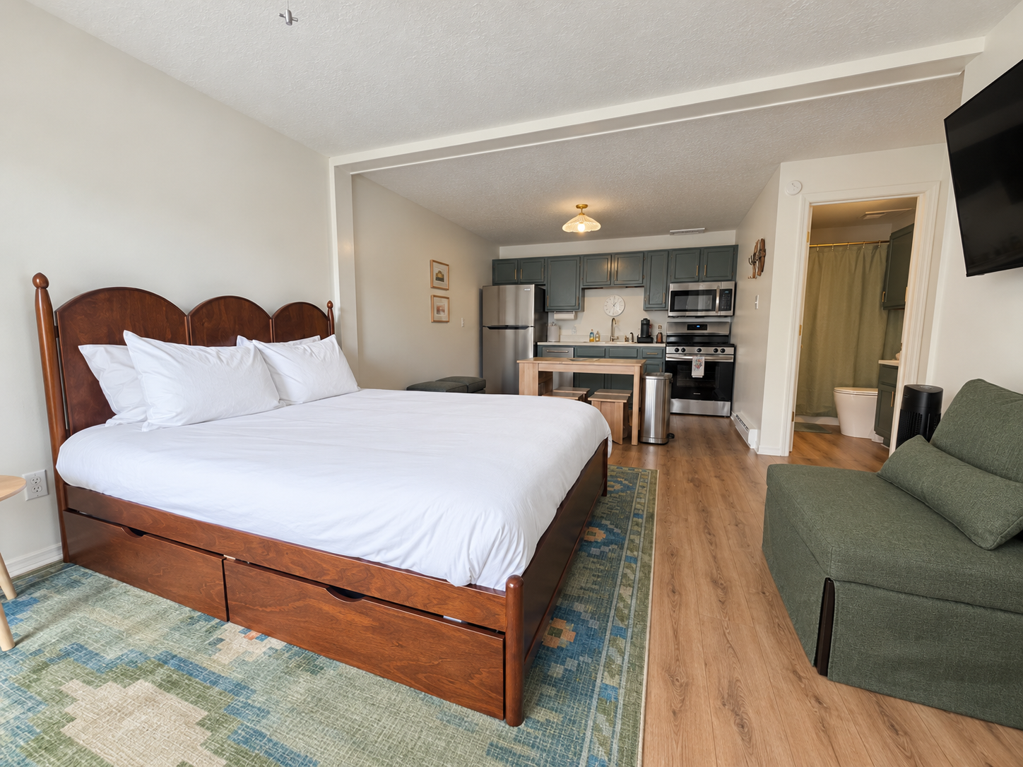 Hotel room with large bed, green armchair, kitchen area with blue cabinets, stainless steel appliances, and a small dining table.