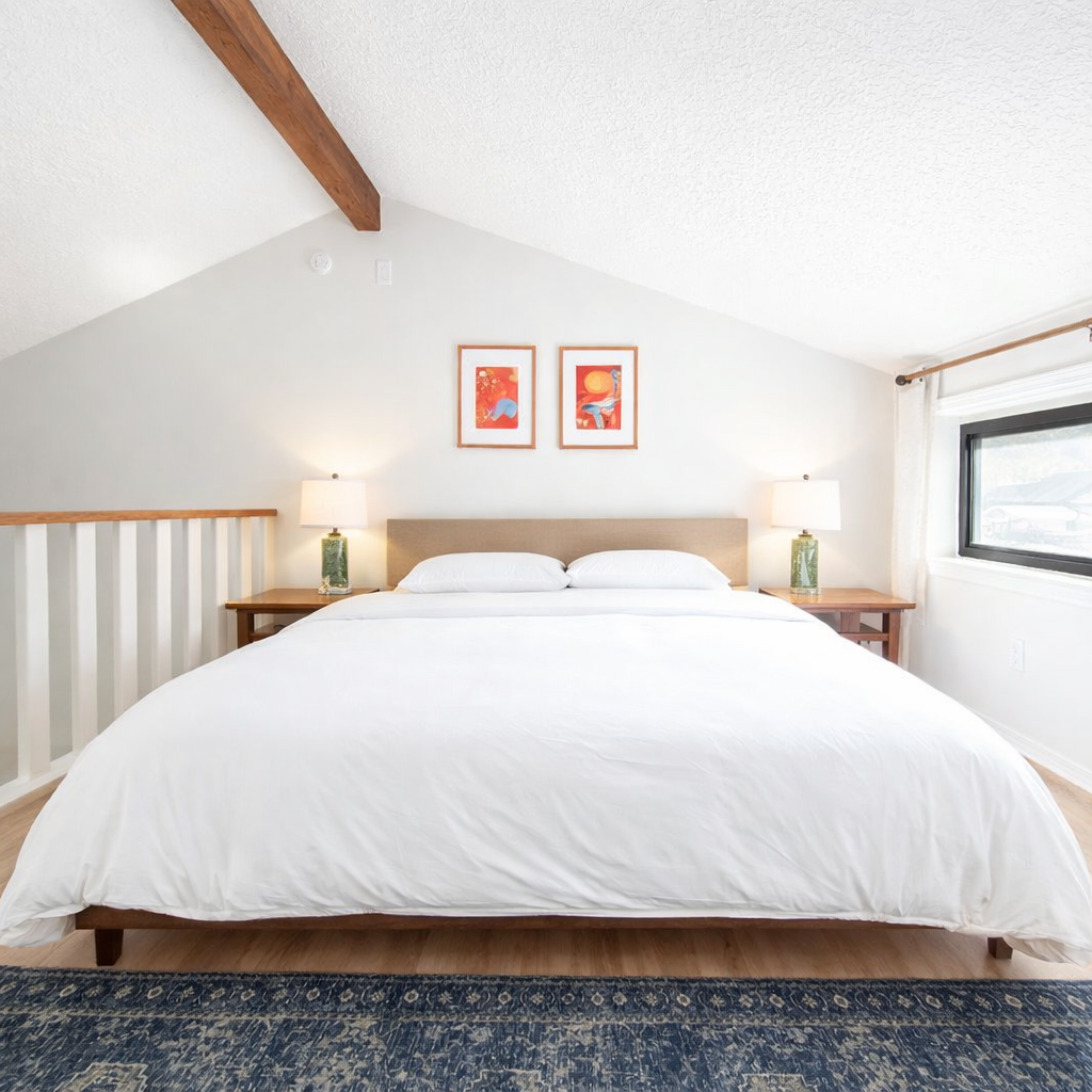 A neatly made bed with white bedding, two side tables with lamps, framed artwork above the bed, and a large window on the right, in a bright bedroom with a sloped ceiling.