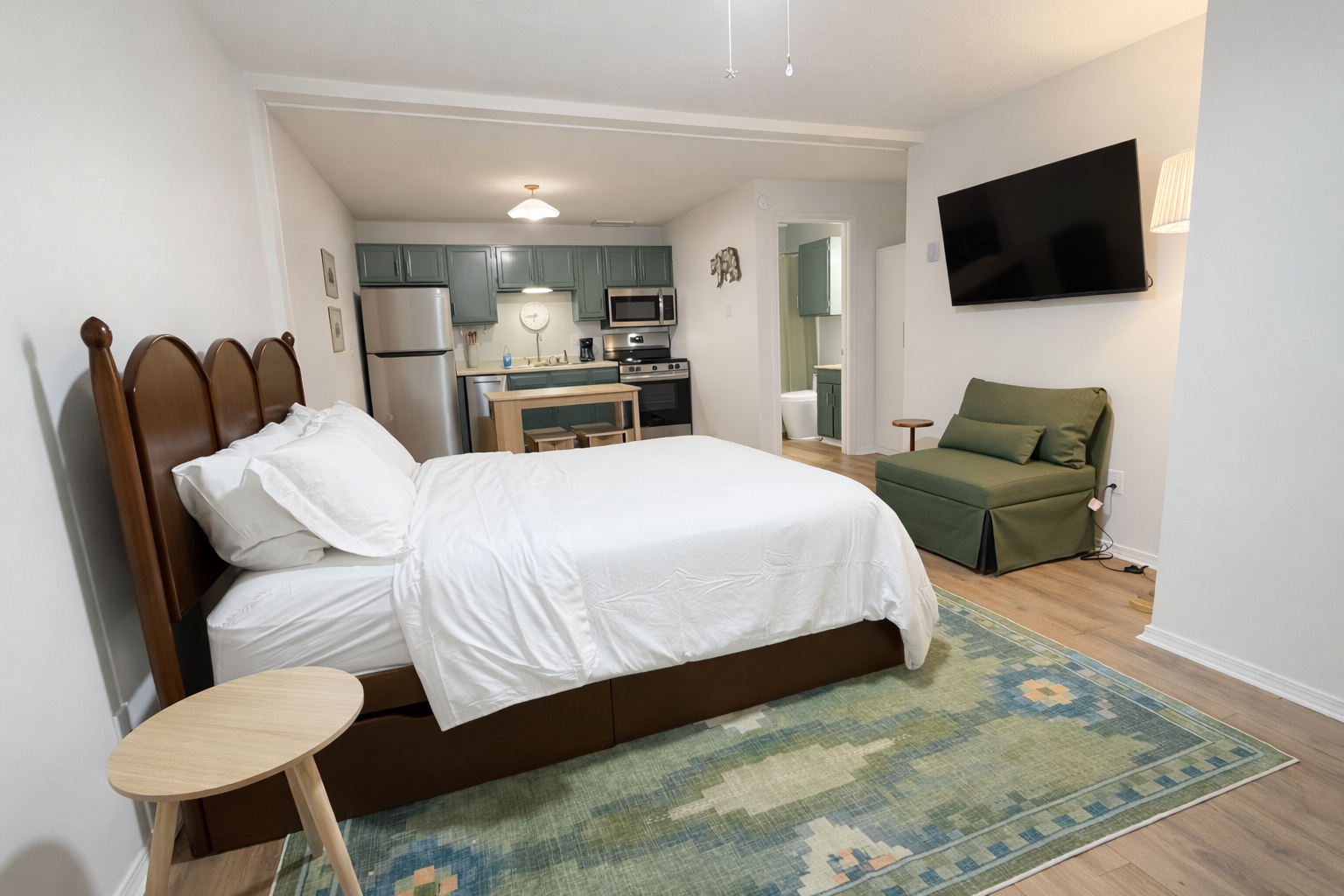 Studio apartment with a bed, small table, kitchen area, green armchair, large wall-mounted TV, and a bathroom in the background.