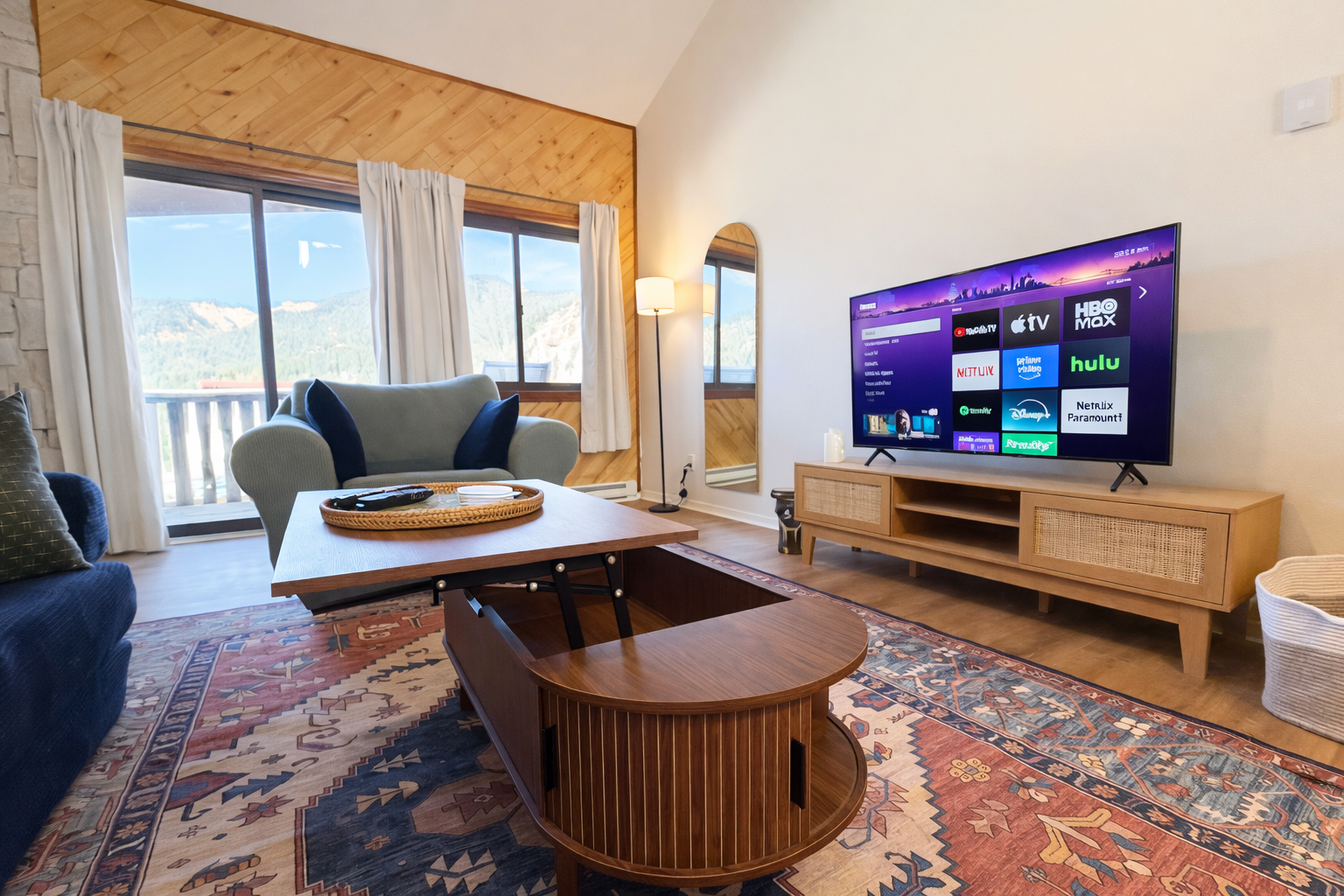 Living room with a large TV displaying streaming app icons, a wooden coffee table, a light green armchair, a dark blue sofa, and sliding glass doors with white curtains leading to a balcony with mountain views.