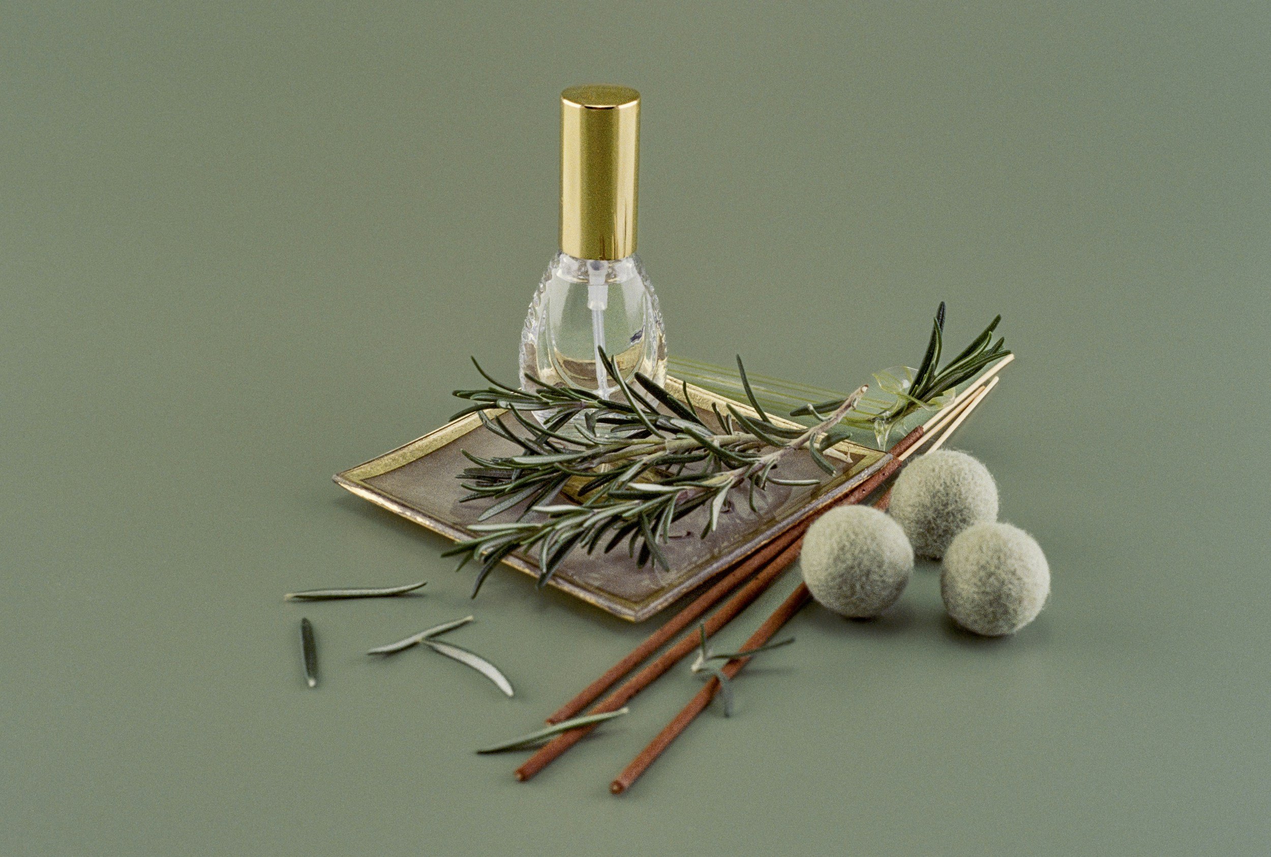 A perfume bottle with a gold cap, a sprig of rosemary, three white bath bombs, a couple of cinnamon sticks, and a glass-like object arranged on a small tray with a textured surface, all on a muted green background.