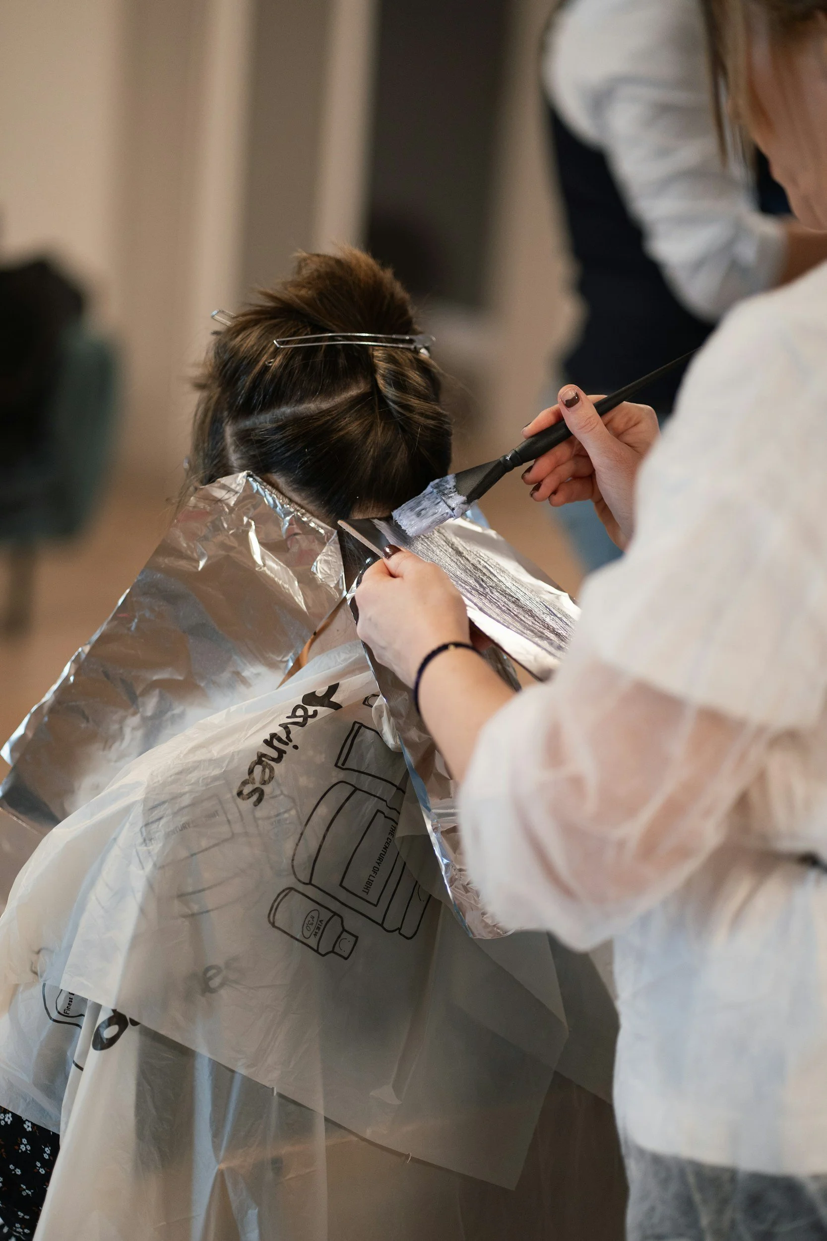 A person getting their hair colored at a salon, with a stylist applying dye using a brush while wearing a white coat, and a client covered with a silver foil cape.
