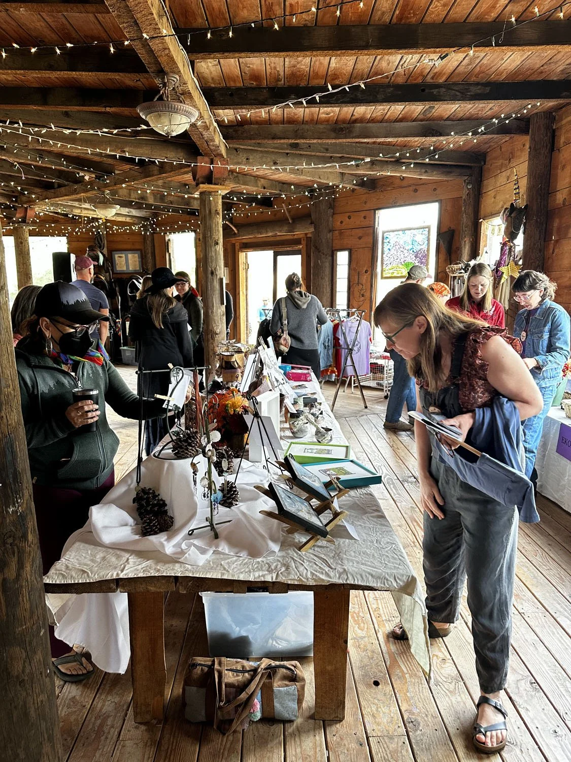 People browsing handmade crafts and artwork displayed on tables inside a rustic wooden building with string lights overhead