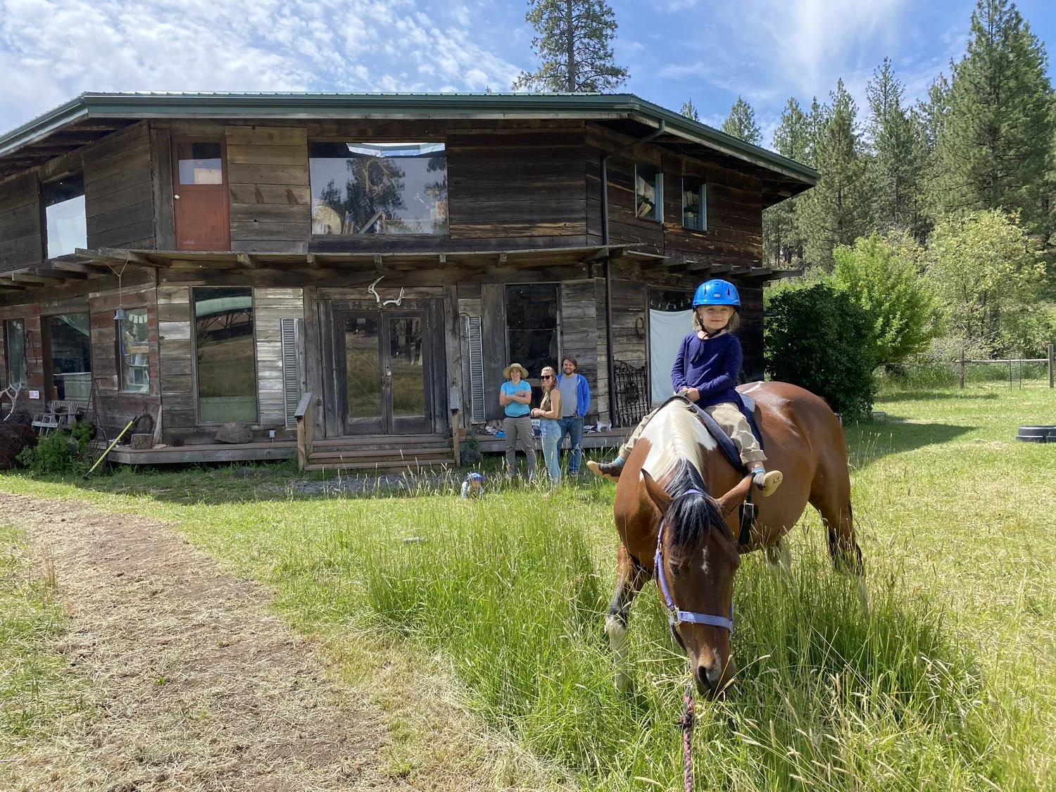 A young girl wearing a blue helmet rides a brown and white horse on a grassy field in front of a rustic two-story wooden house surrounded by trees.