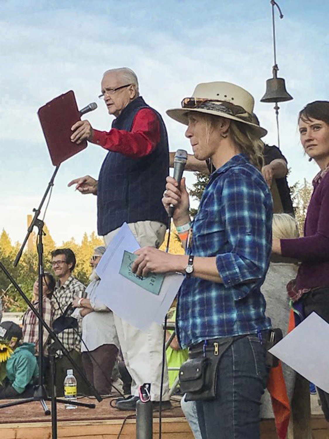 People speaking at an outdoor event, with one man holding a microphone and reading from a paper, and another person wearing a hat also holding a microphone. Others are standing nearby, and a diverse crowd is in the background.