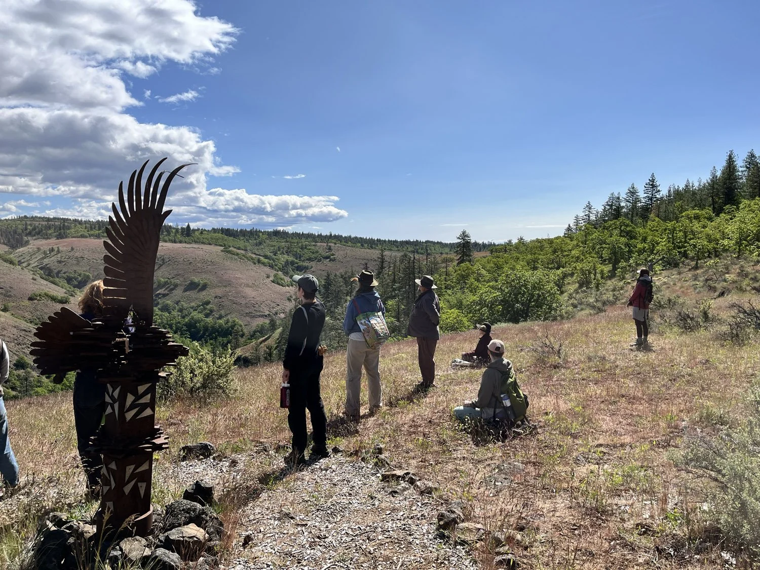 A group of hikers with backpacks and hats standing and sitting on a hillside with sparse vegetation, trees in the background, and a sculpture of a feather on the left side, under a partly cloudy sky.