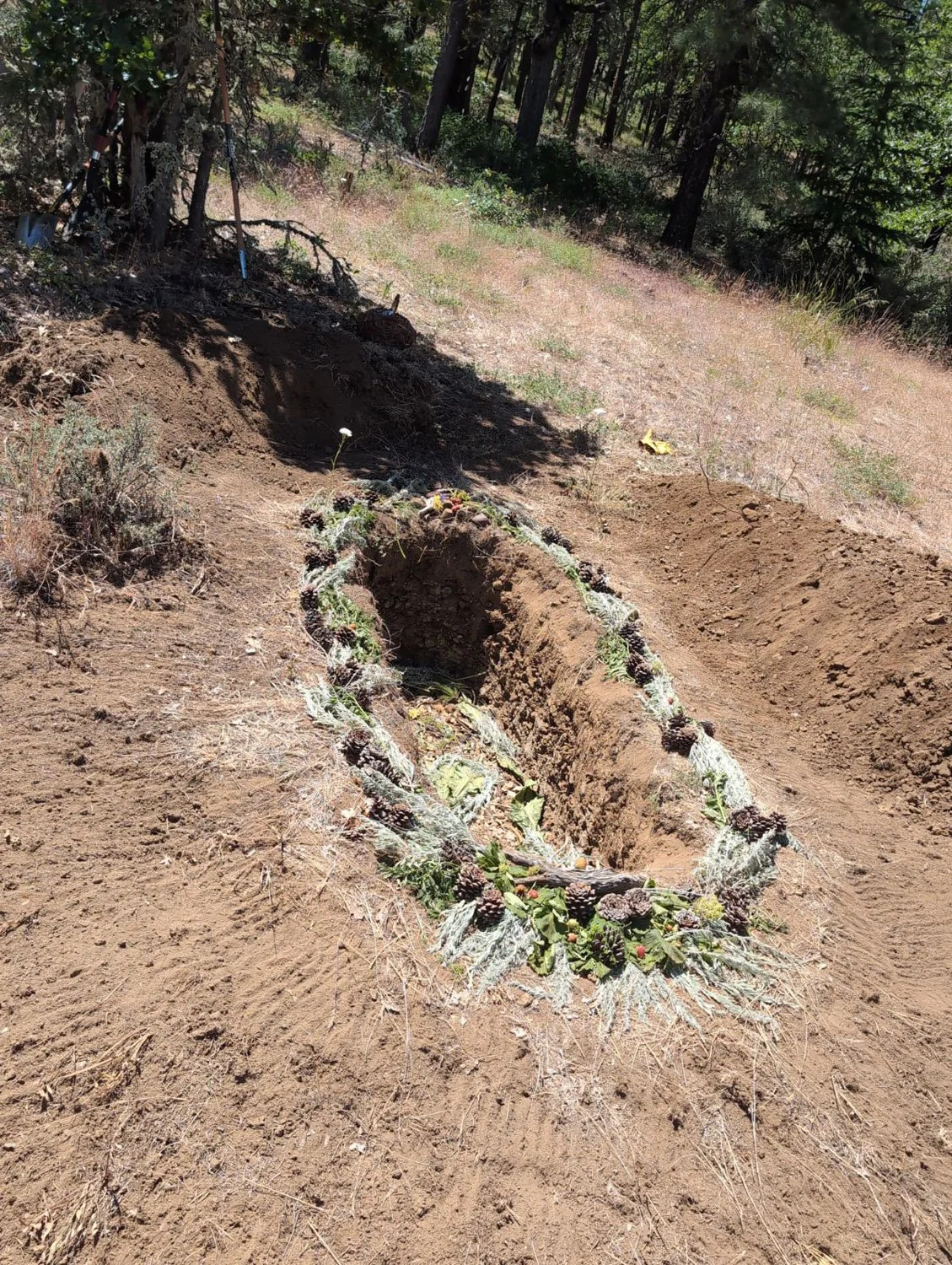 A newly dug grave in a wooded outdoor area, decorated with pinecones, leaves, and natural ornaments.