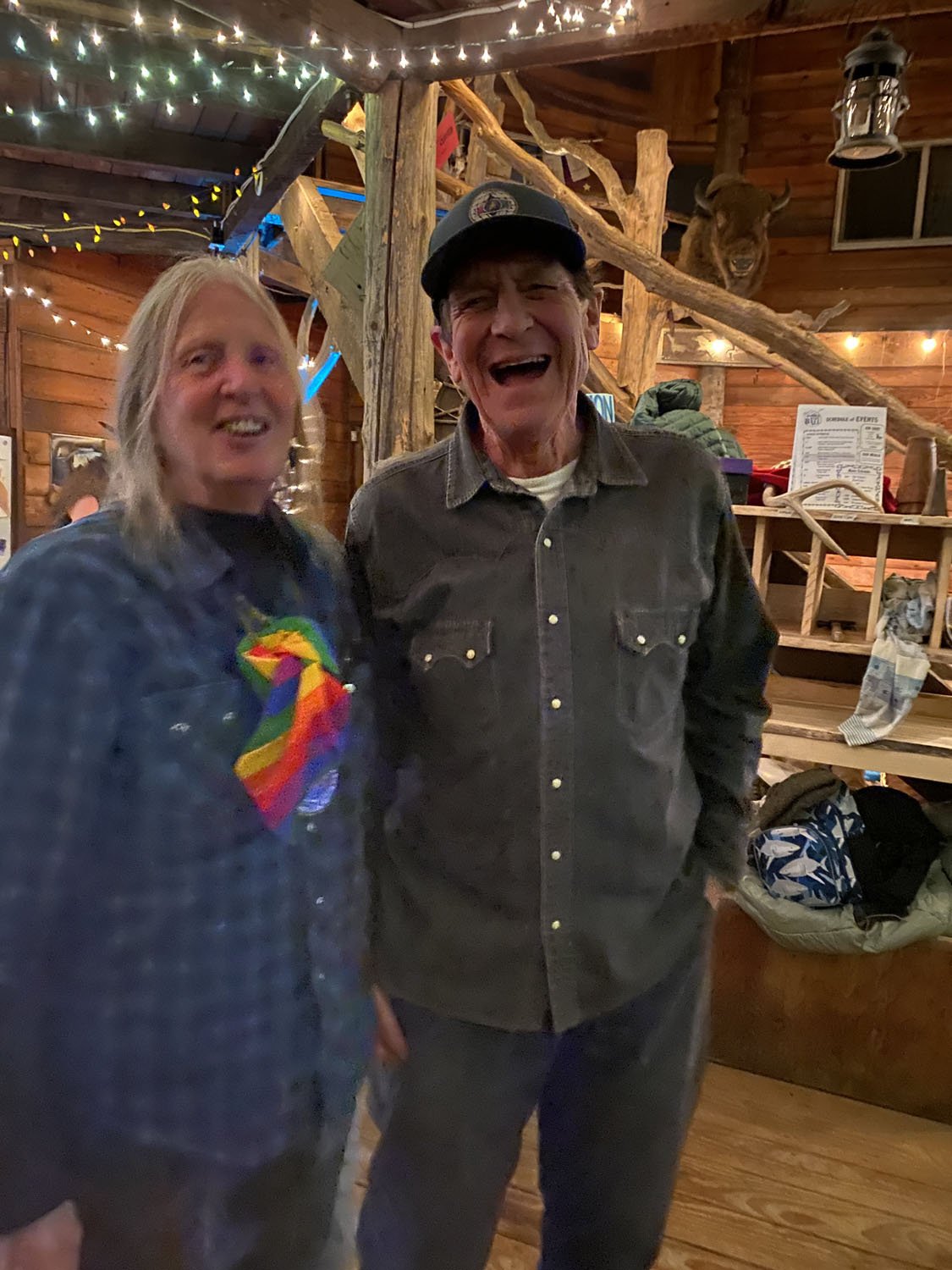Two people, a woman with shoulder-length blonde hair and a man wearing a black cap, are smiling and standing close together inside a rustic wooden lodge decorated with string lights, a mounted animal head, and various lodge-related items.