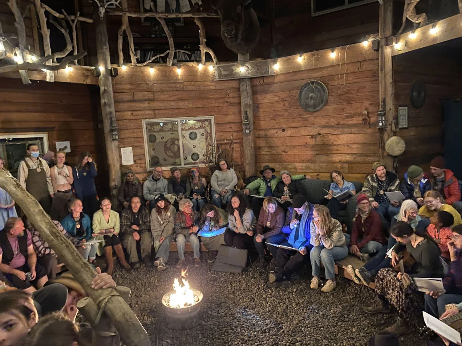 A group of people gathered around a campfire inside a wooden cabin with rustic decor, some seated on benches and others standing, engaging in a storytelling or discussion activity.