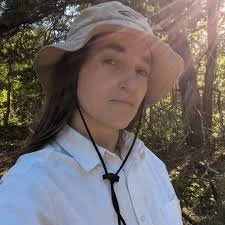 Woman wearing a wide-brimmed hat and white shirt outdoors among trees, sunlight in the background.