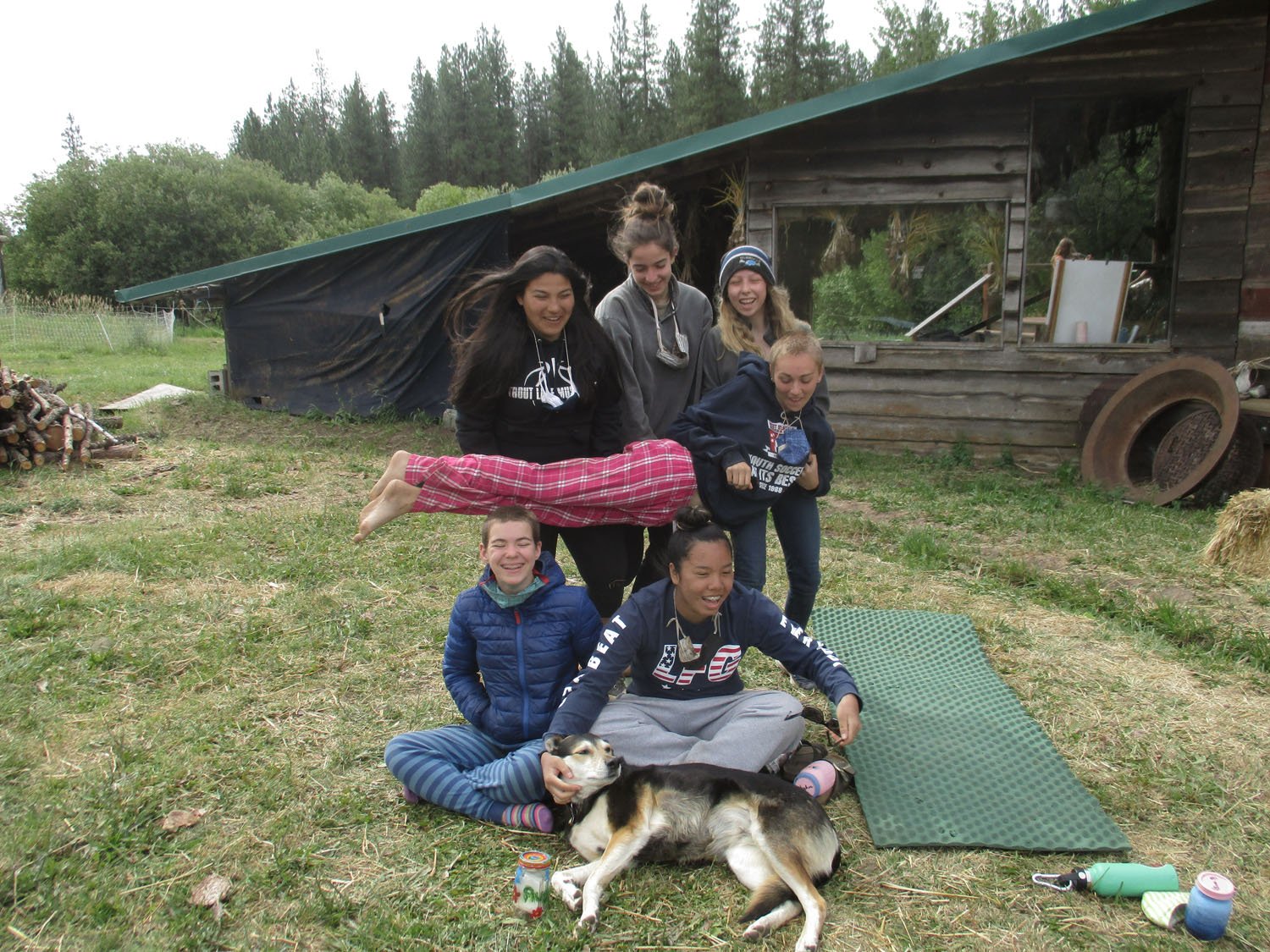 Group of young women and a dog outside near a wooden shed, with some toys and a mat on the ground.