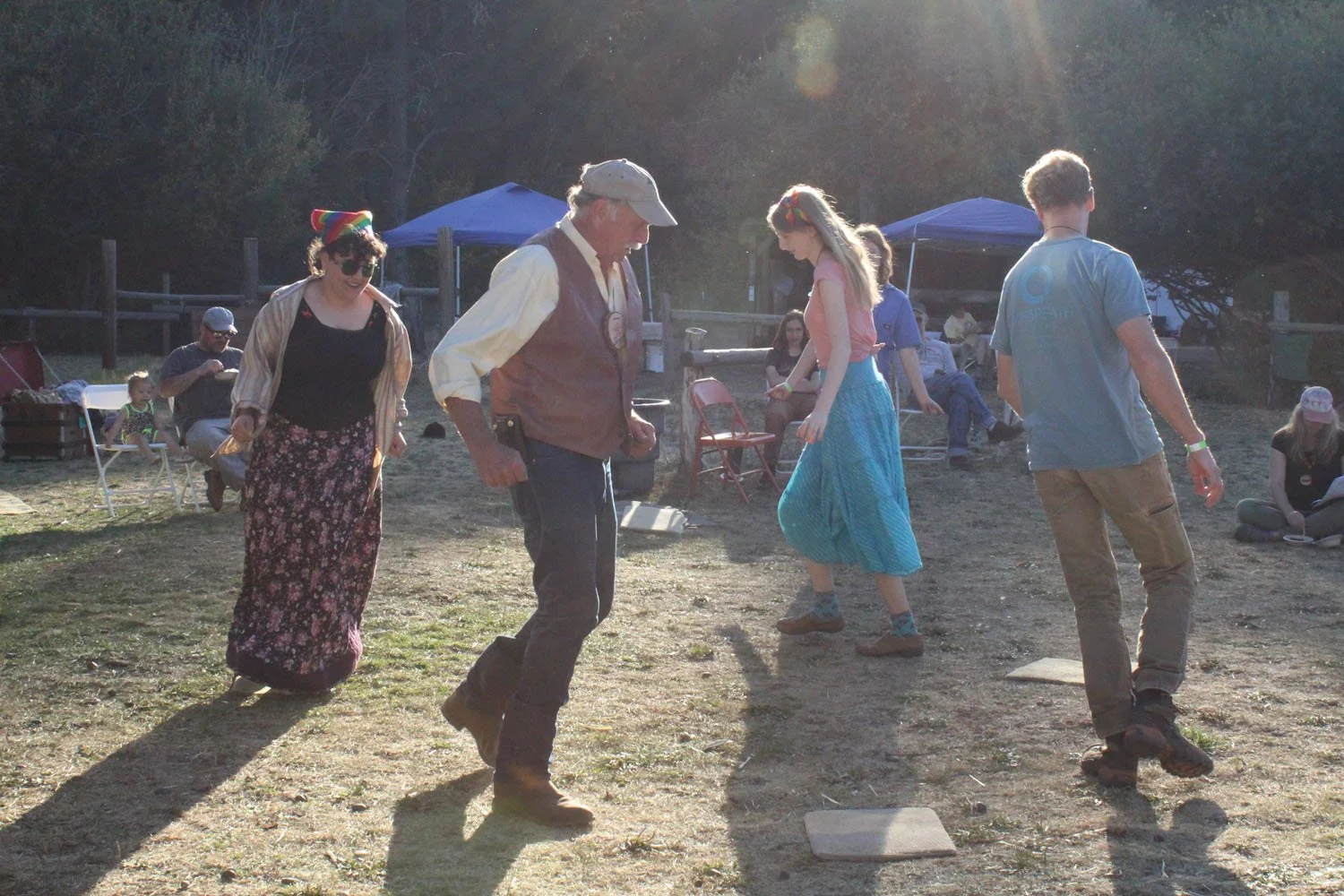 People playing a game outdoors on a dirt field during the evening, with tents and other people sitting and standing in the background.