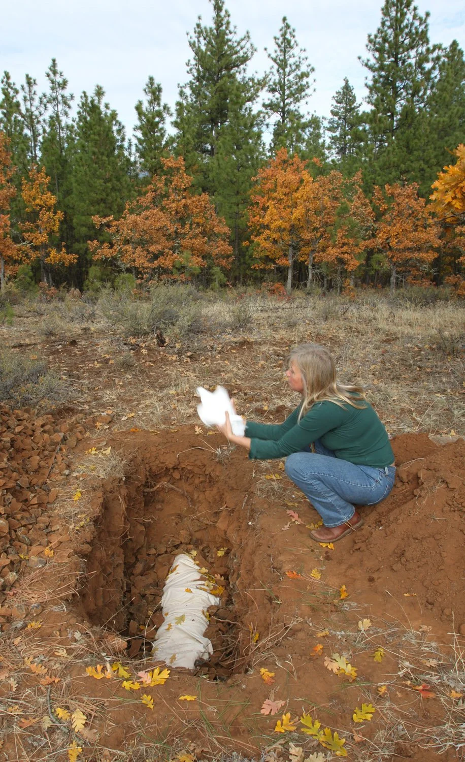 A woman crouching near a trench in an autumn forest, holding a white object and looking into the trench with fall leaves scattered around.