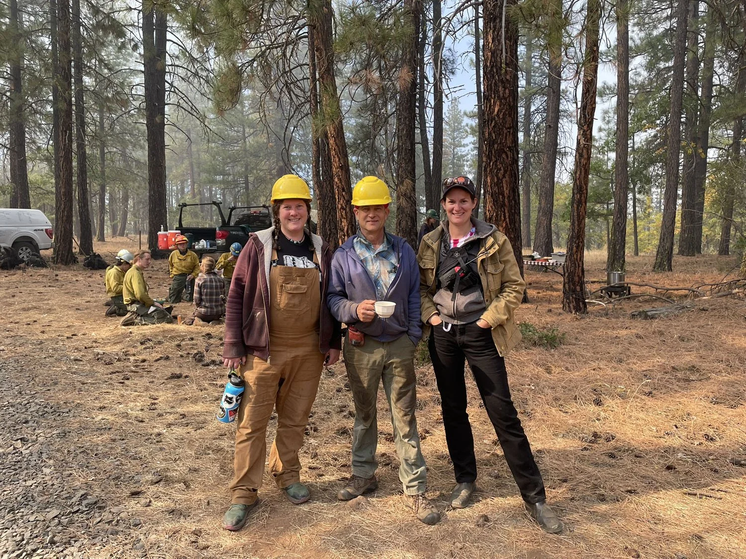 Three women in forestry work gear, including yellow helmets, standing in a forest with other workers sitting on the ground and equipment in the background.