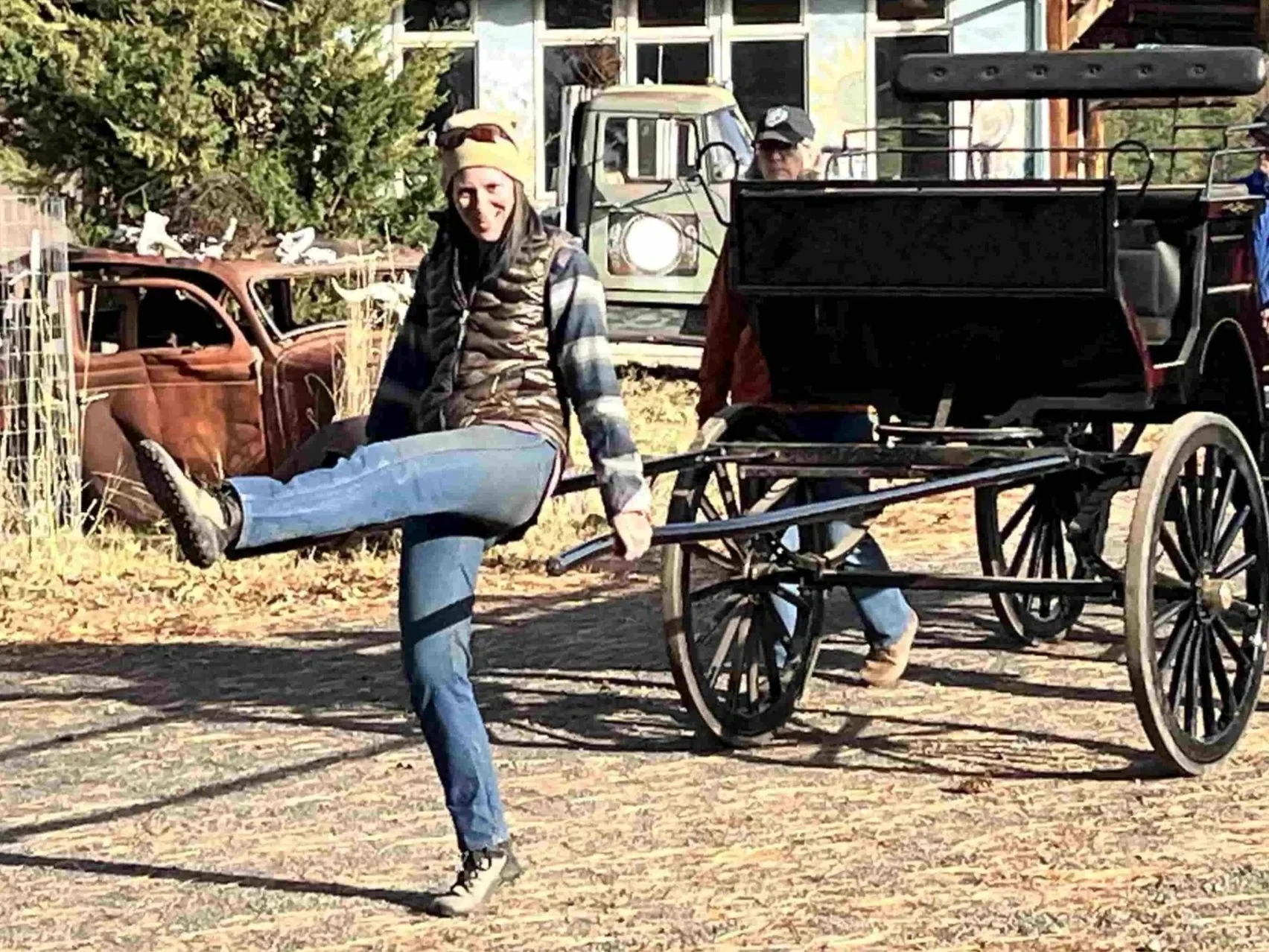 Woman in casual clothing and a cap kicking her leg up while standing next to a horse-drawn carriage with two wheels, with two men in the background and old cars and trees in the background.
