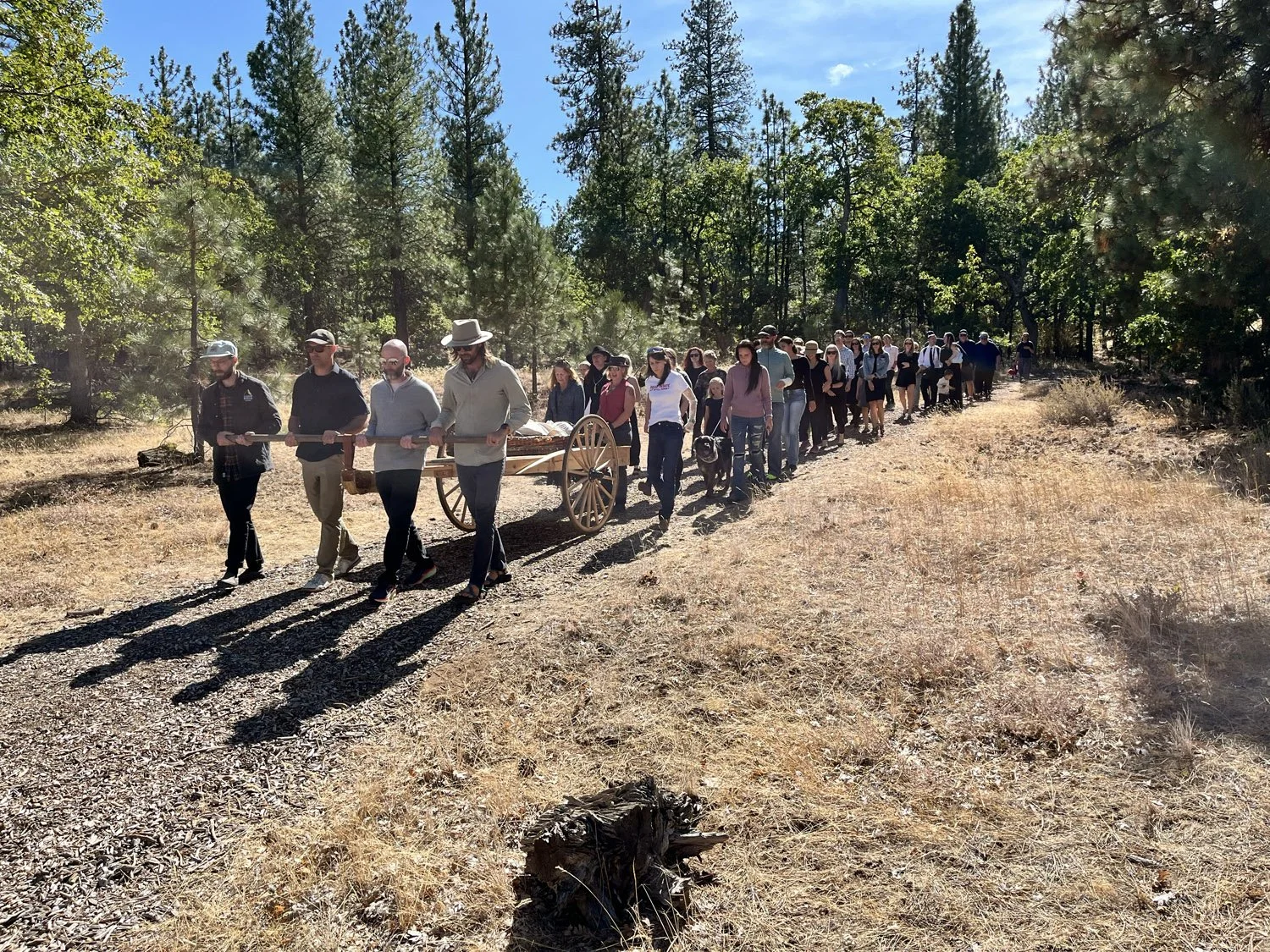 A group of people walking on a dirt trail in a forested area, some wearing hats, with one person at the front pushing a wooden cart.