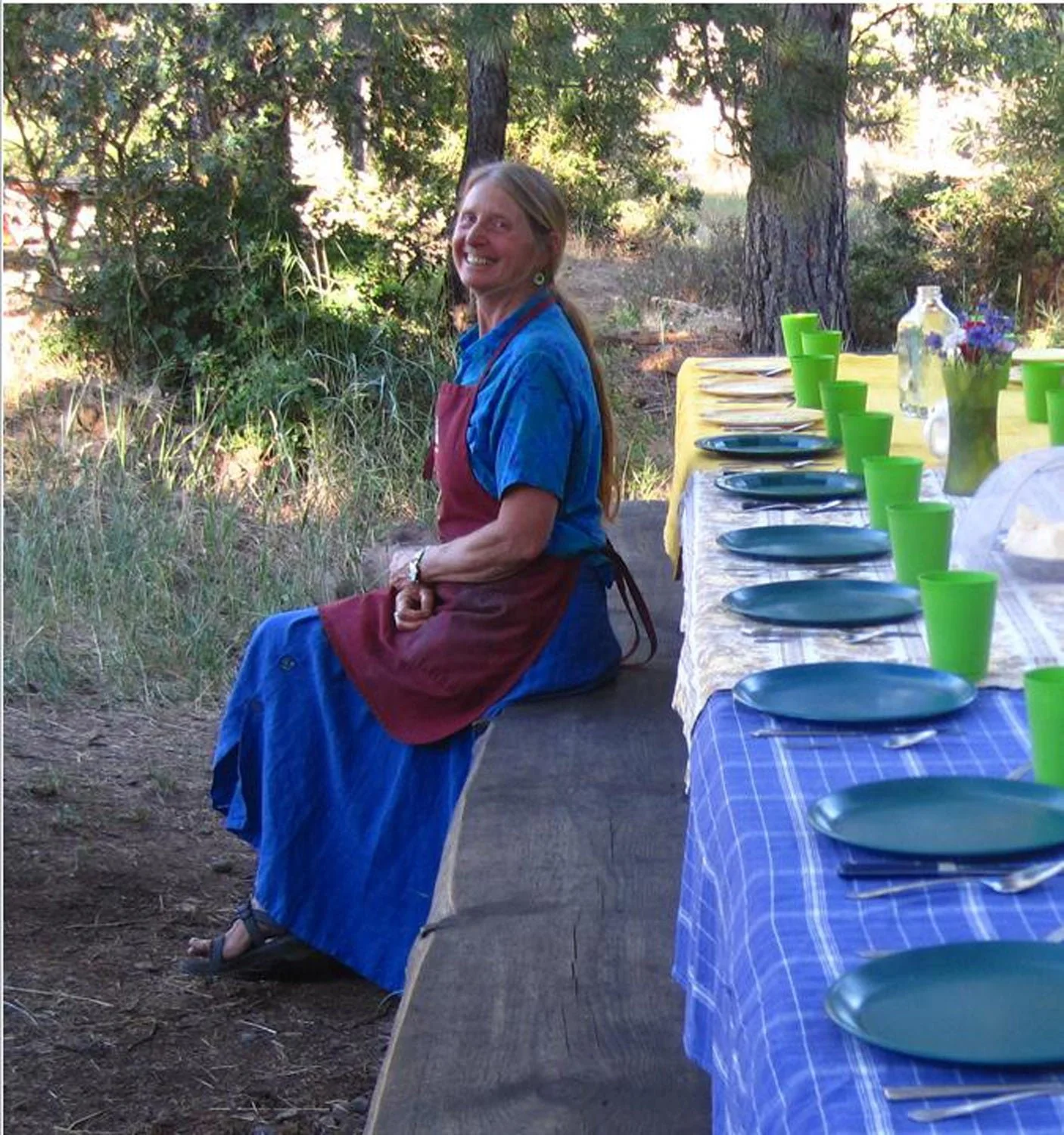 A woman sitting outdoors at a rustic table set for a meal, with green cups, plates, and a vase with flowers, surrounded by trees and nature.