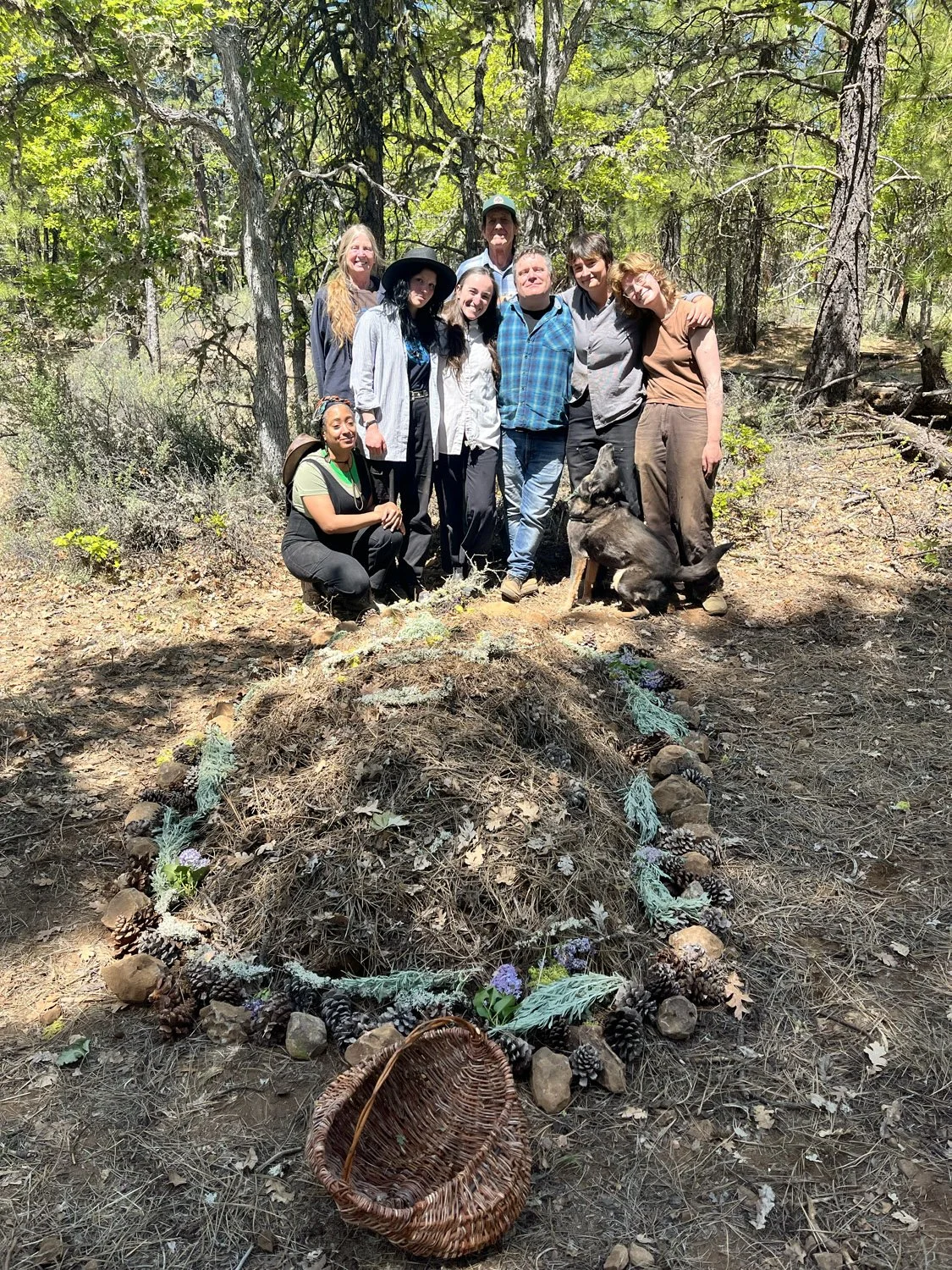 A group of nine people and a dog standing together in a forest, behind a decorated mound with pinecones, leaves, and flowers, and an empty woven basket in front.
