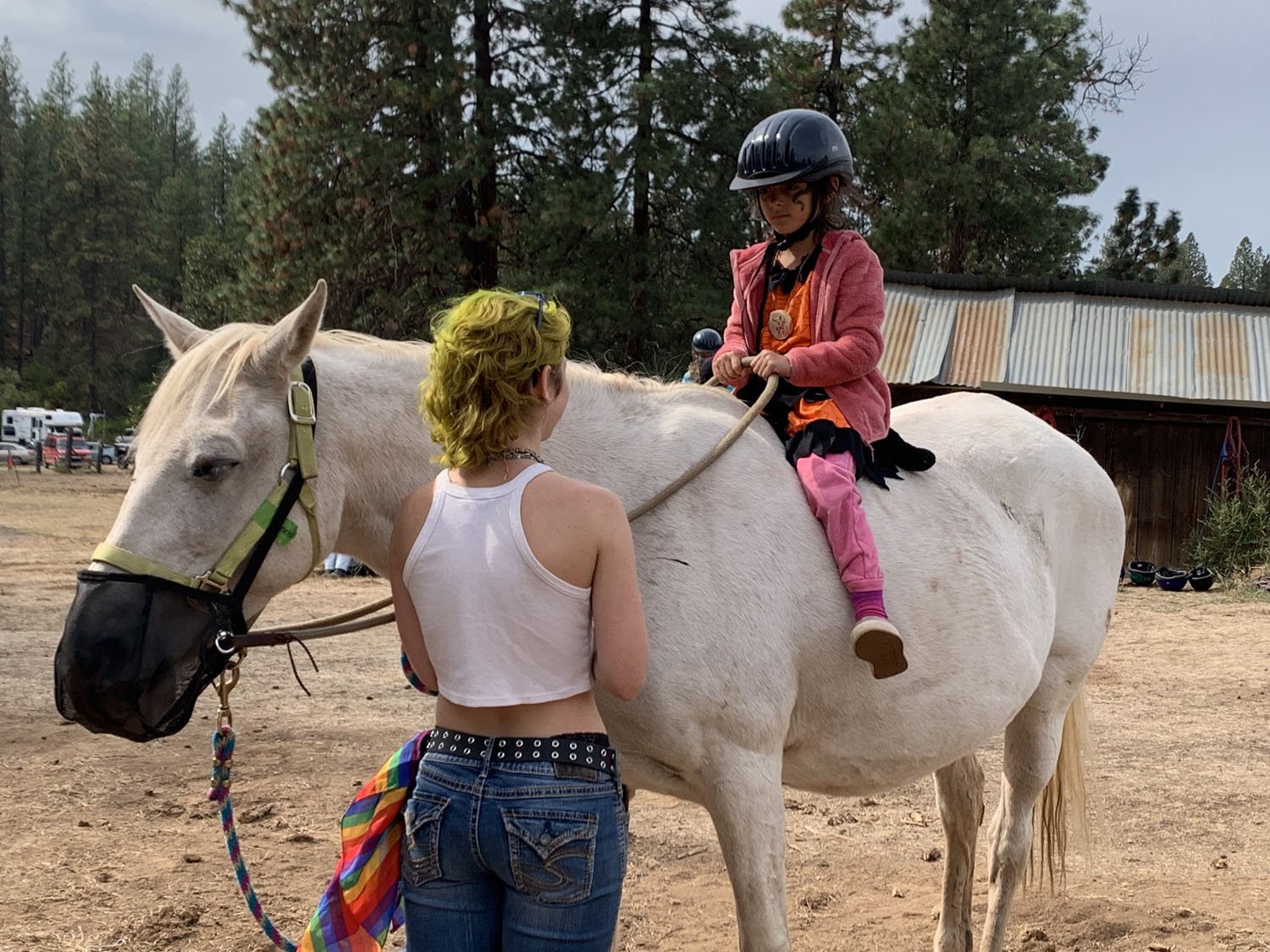 A young girl wearing a pink hoodie and black helmet sits on a white horse, while a woman with short yellow hair and a white sleeveless shirt stands nearby holding the horse's reins. The setting is outdoors with trees and a rustic building in the background.