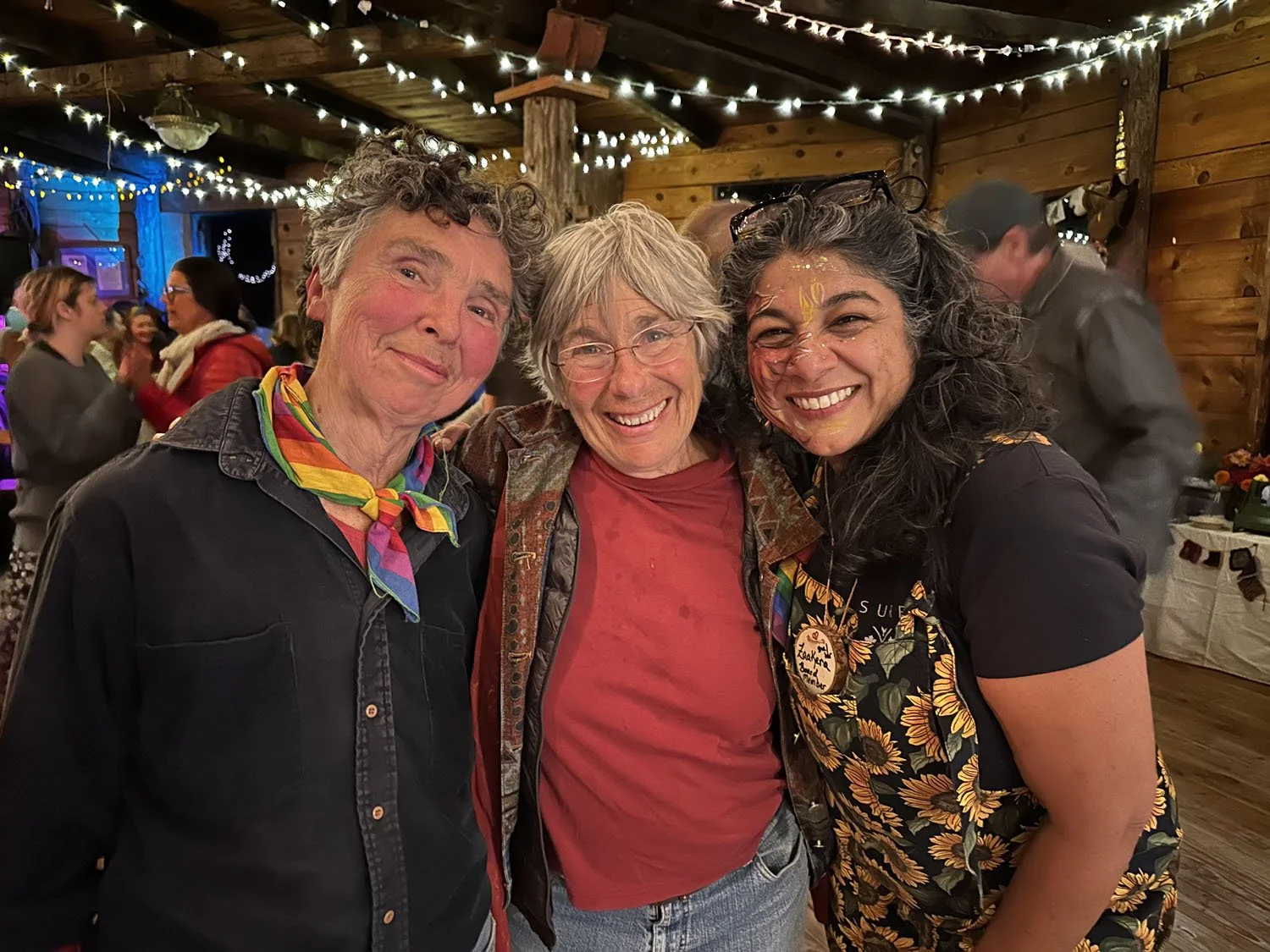 Three women smiling at a social gathering or party inside a rustic venue with wooden walls and string lights. One woman has gray hair, glasses, and wears a colorful scarf. The woman on the right has dark curly hair, glasses on her head, and face paint.