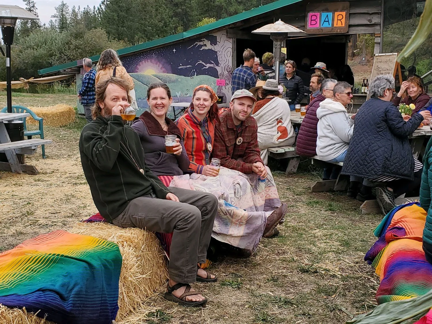 People sitting and standing outside at a gathering, some drinking, with a bar in the background, and hay bales and a colorful rainbow blanket in the foreground.