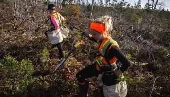 Two Scott and Stewart tree planters climb through a rugged feild with sacks of trees over each shoulder and tree planting tools in their hands.