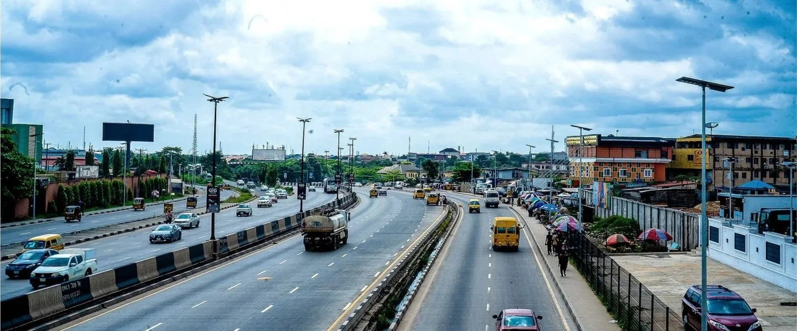 A busy multi-lane highway with cars, trucks, and buses, and a sidewalk with pedestrians using umbrellas under cloudy skies.