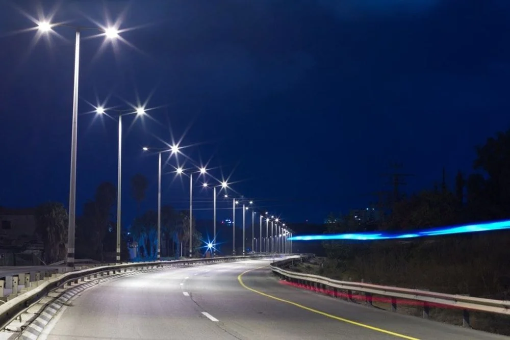 Empty highway at night with streetlights, guardrails, and a faint trail of light from a moving vehicle.